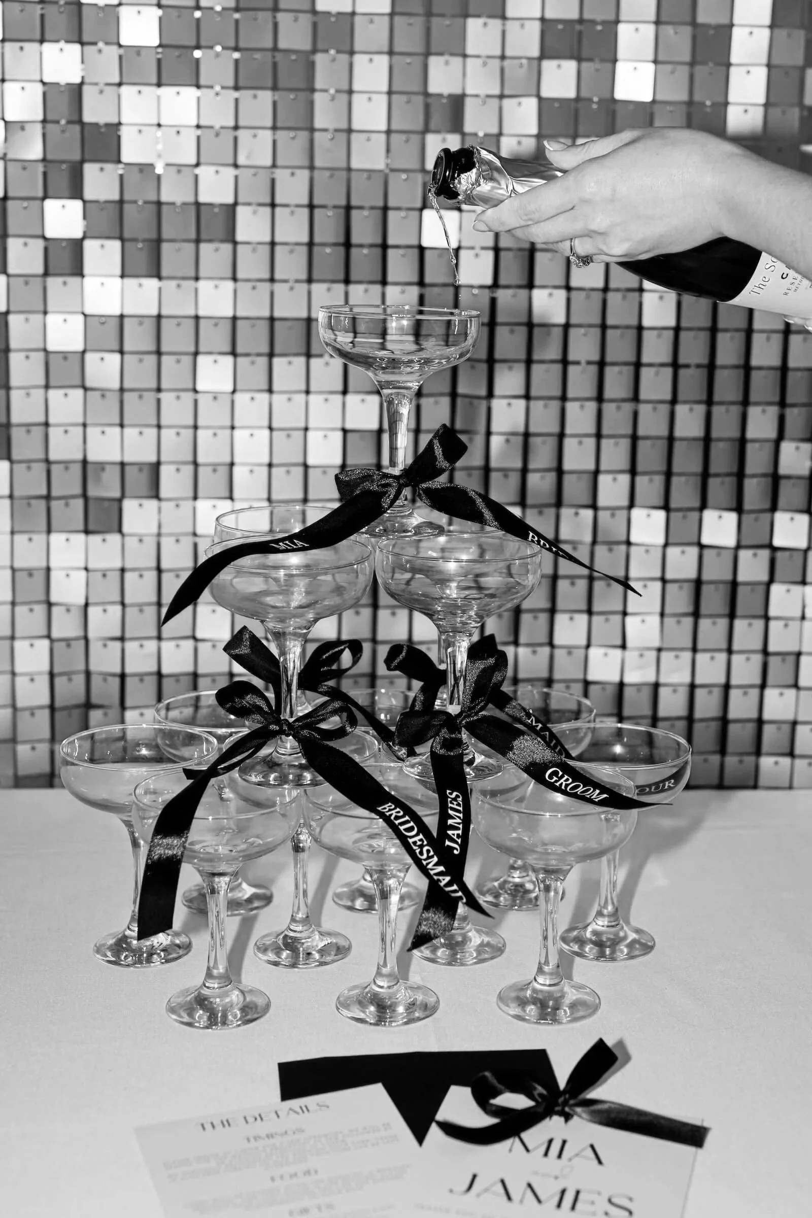 Stacked coupe glasses with black ribbons and bows, a champagne bottle being poured into the top glass, with a patterned background and wedding details on a paper at the front.