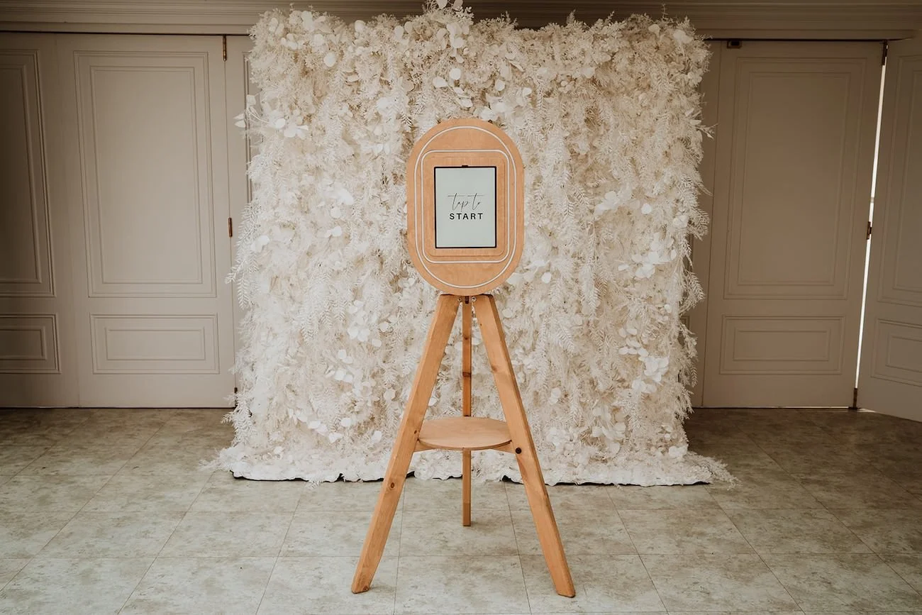A wooden photo booth with a screen displaying "tap to start" is set against a white floral backdrop in an indoor setting.