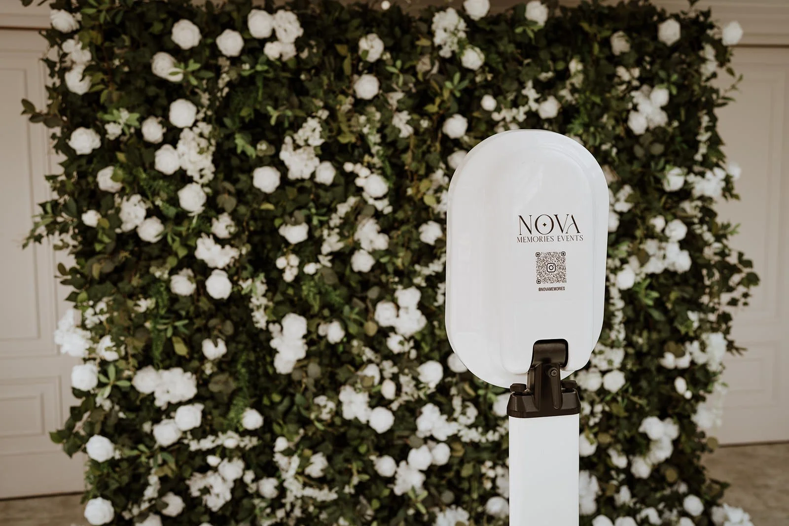 A white photo booth with a logo that reads 'NOVA Memories Events' and a QR code, set against a backdrop of white flowers and green leaves.