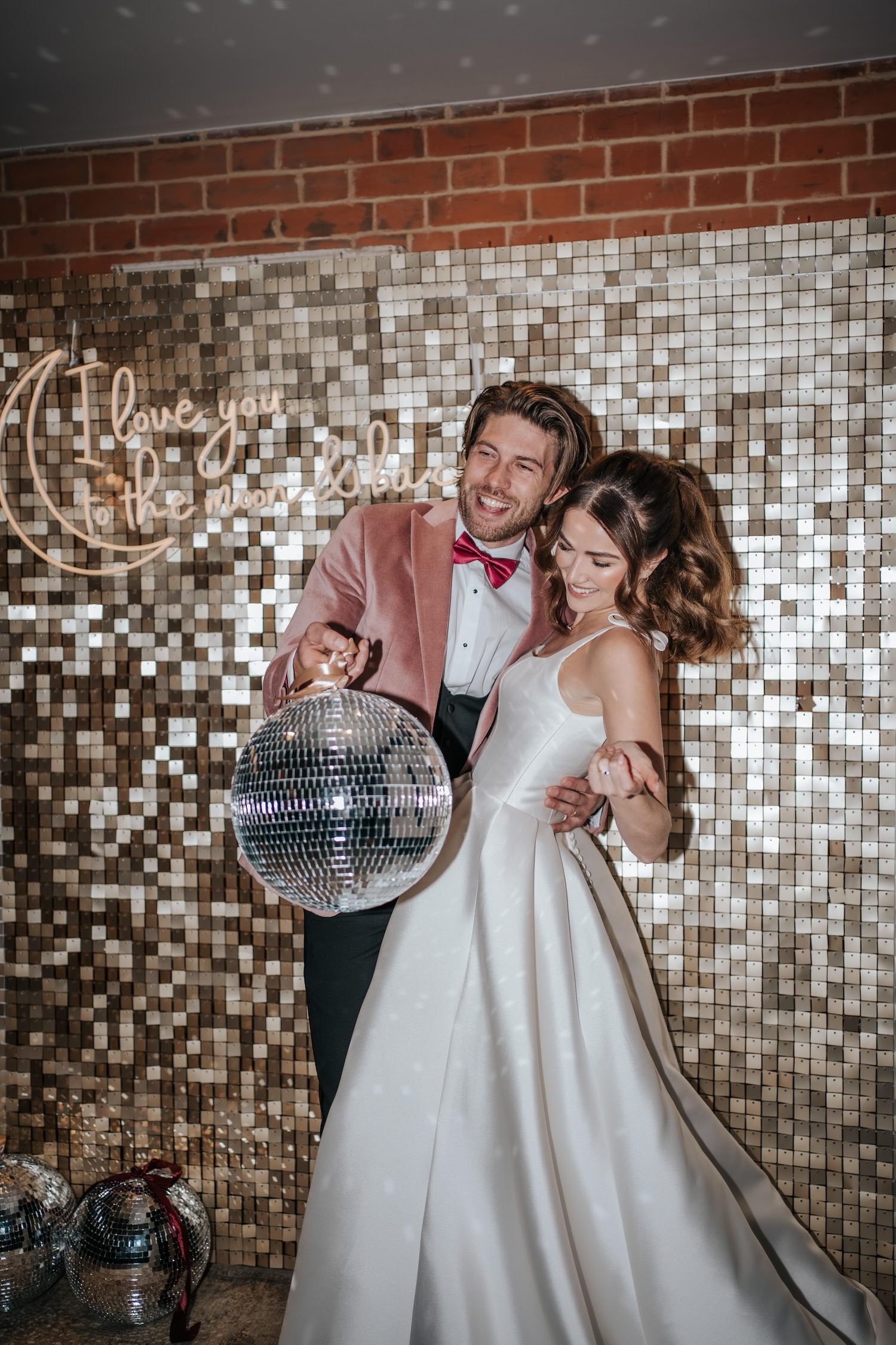 A couple dressed in wedding attire dancing with a disco ball. The bride is wearing a white gown, and the groom is wearing a pink suit with a red bow tie. Behind them is a glittery, tiled wall with a neon sign that reads, 'I love you to the moon and b