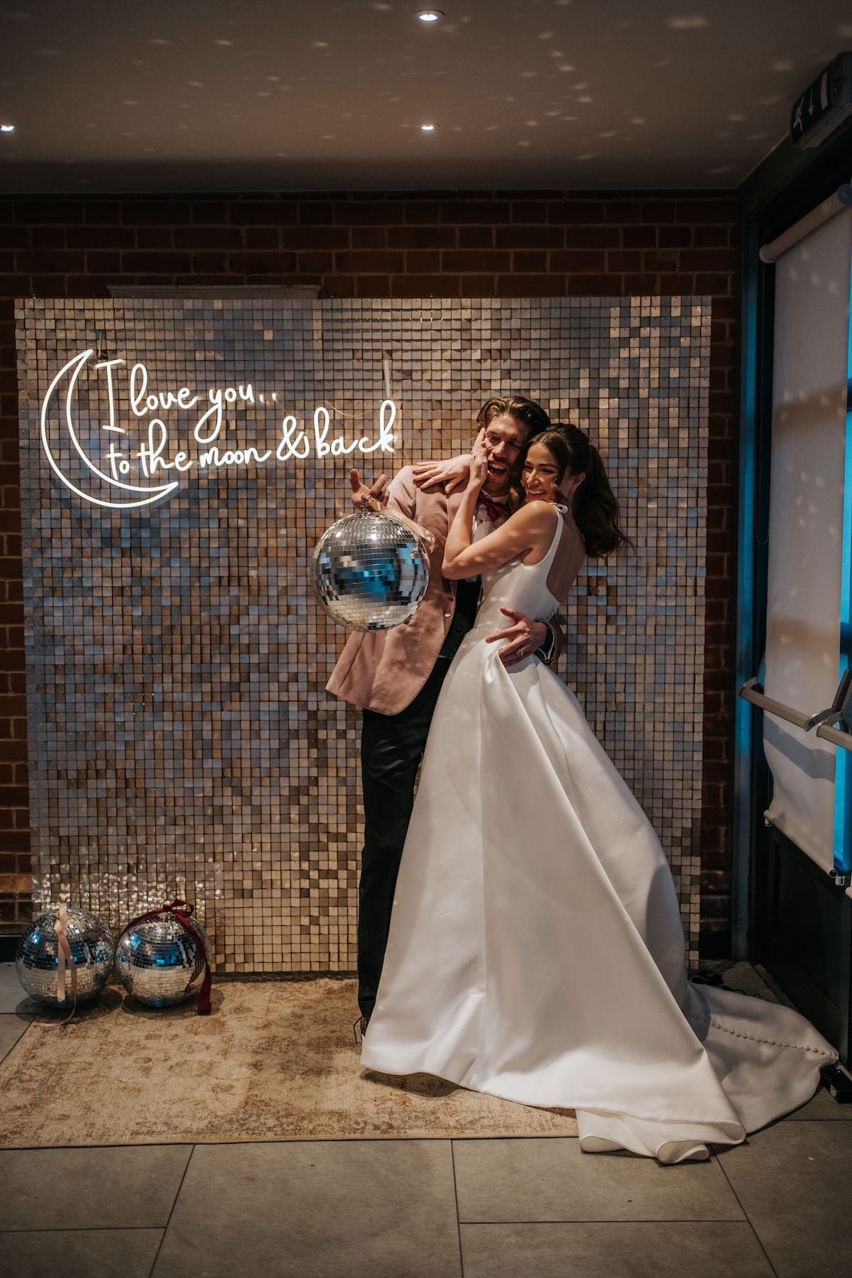 A bride and groom dancing together at their wedding reception, with a disco ball and a sign that says 'I love you to the moon and back' in the background.