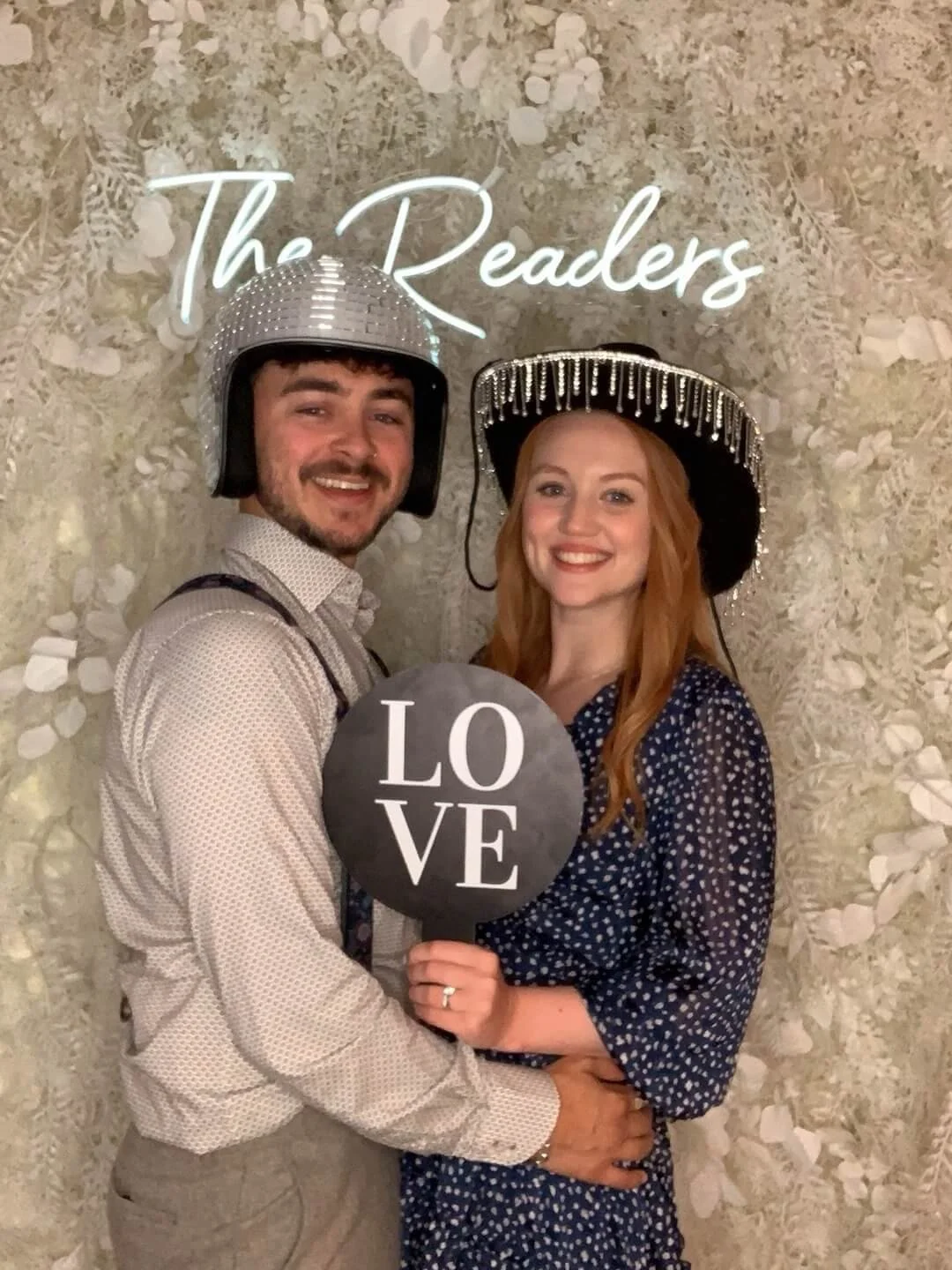 A smiling man and woman dressed festively with shiny hats, holding a black circle shape sign that says "LOVE". They are standing in front of a floral background with neon lettering that reads "The Readers."
