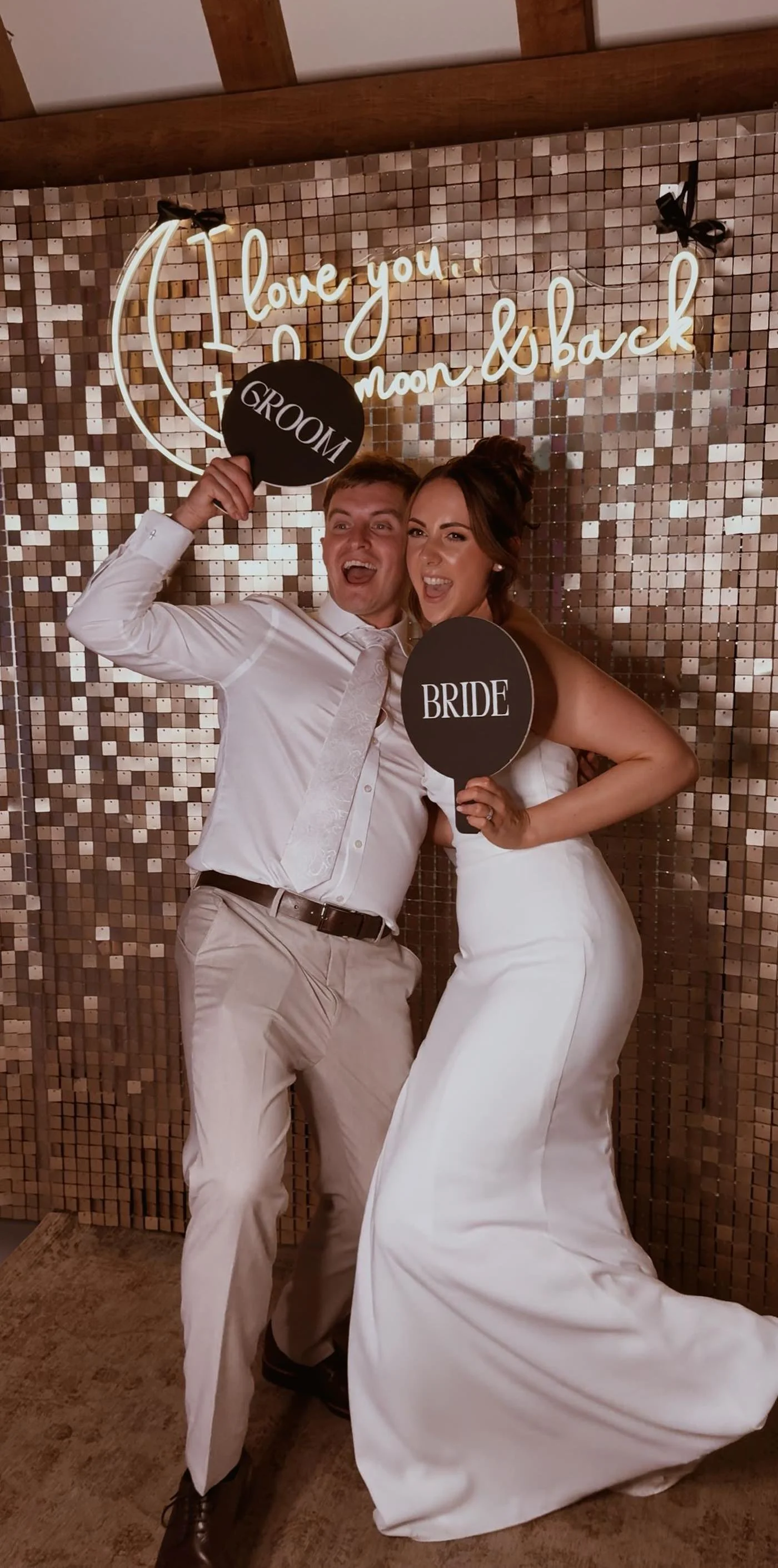 A bride and groom celebrating at their wedding, holding signs that say 'GROOM' and 'BRIDE', standing in front of a decorative wall with a neon sign that says 'Love you to the moon & back'.