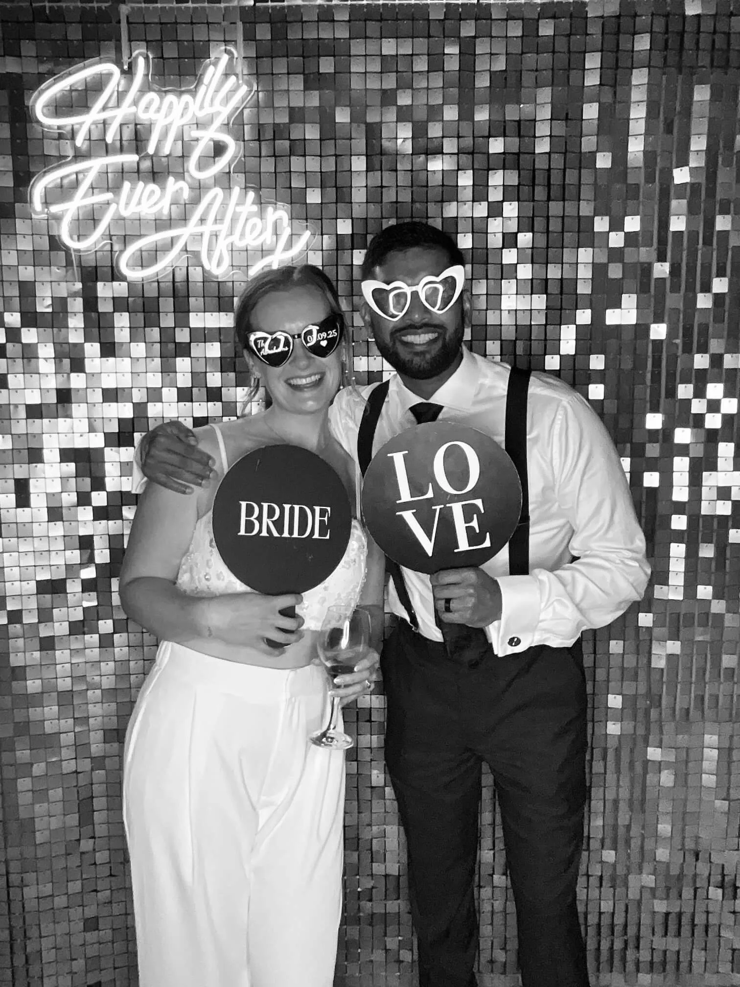 A bride and groom at a wedding, smiling and holding glasses of wine, standing in front of a tiled wall with a neon sign that says 'Happily Ever After'. The bride is wearing a white dress and sunglasses, while the groom is dressed in a shirt, tie, sus