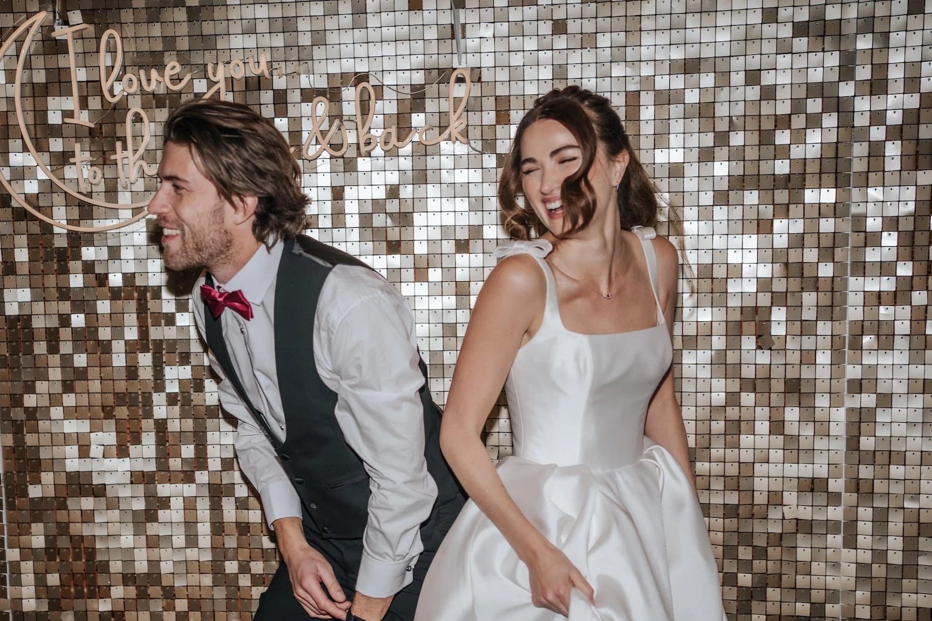 A smiling bride and groom standing back-to-back in front of a decorative metallic wall with a sign that reads 'love you to the back' in cursive script. The groom is wearing a white shirt with a black vest and red bow tie, and the bride is in a white wedding dress.