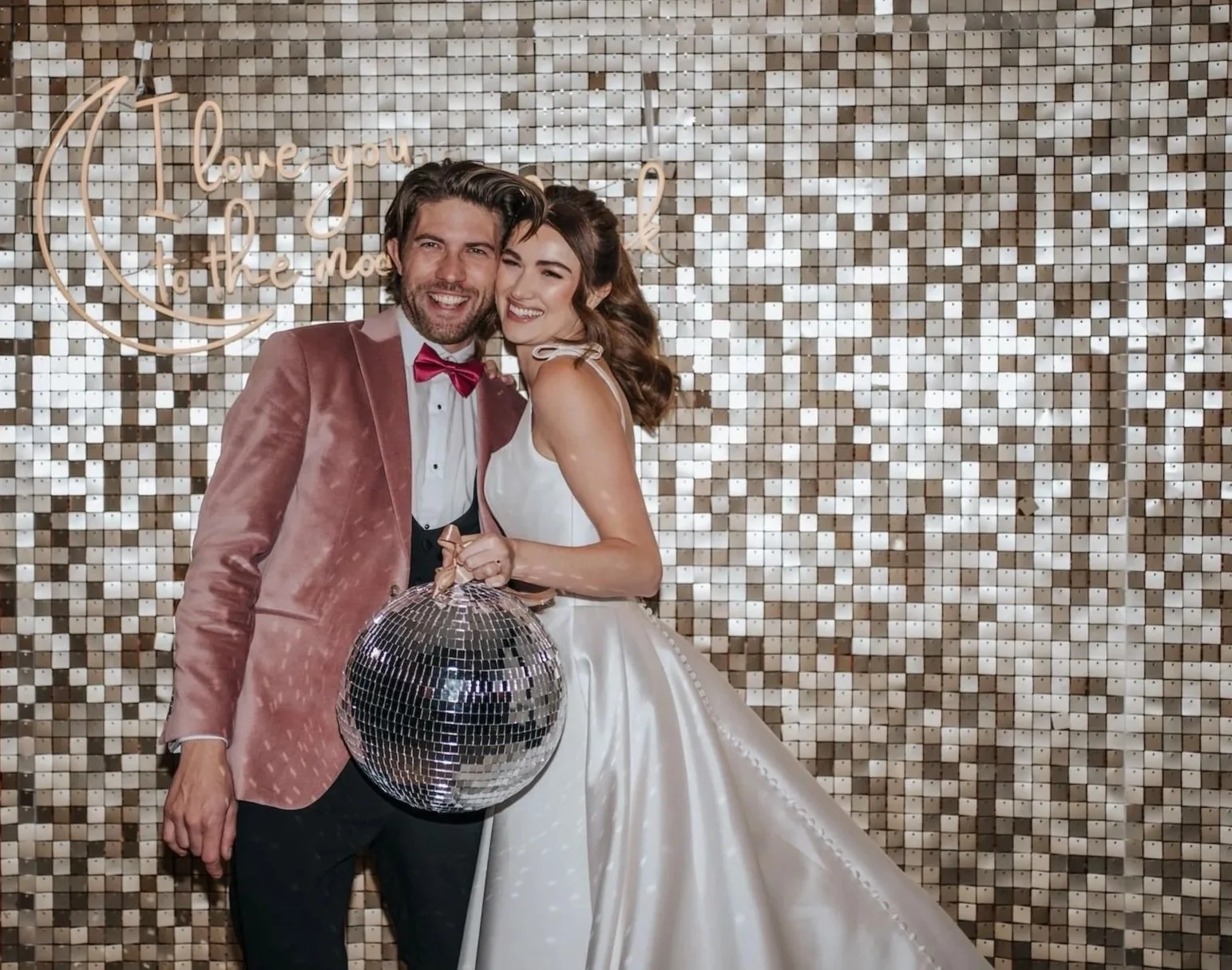 A couple dressed in formal wedding attire, standing in front of a shimmering gold mosaic wall, holding a disco ball with Christmas lights reflected on it, smiling and embracing.