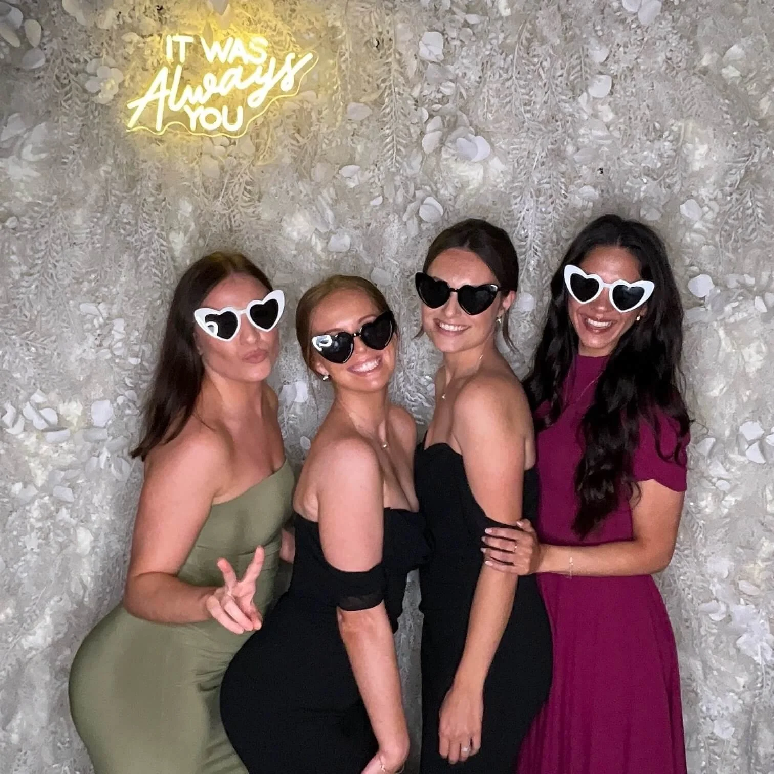 Four women in elegant dresses and large heart-shaped sunglasses pose for a photo against a textured floral wall. The woman on the left wears a green strapless dress and makes a peace sign with her hand. The second woman from the left wears a black st