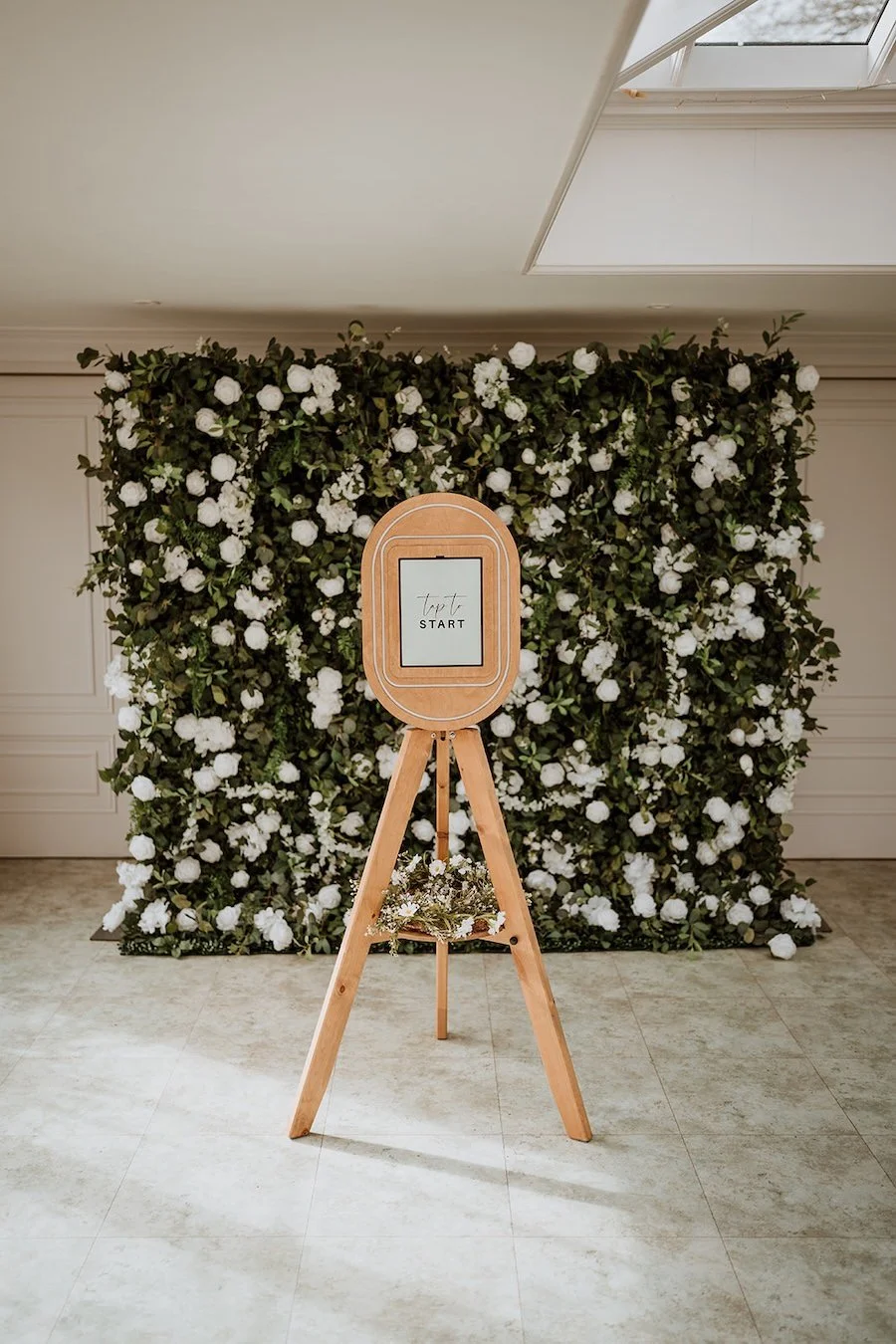 A wooden photo booth digital screen displaying 'START' and the word 'tapt' above it, set in front of a lush floral backdrop of white flowers and green foliage, near a skylight in a room with tiled flooring.