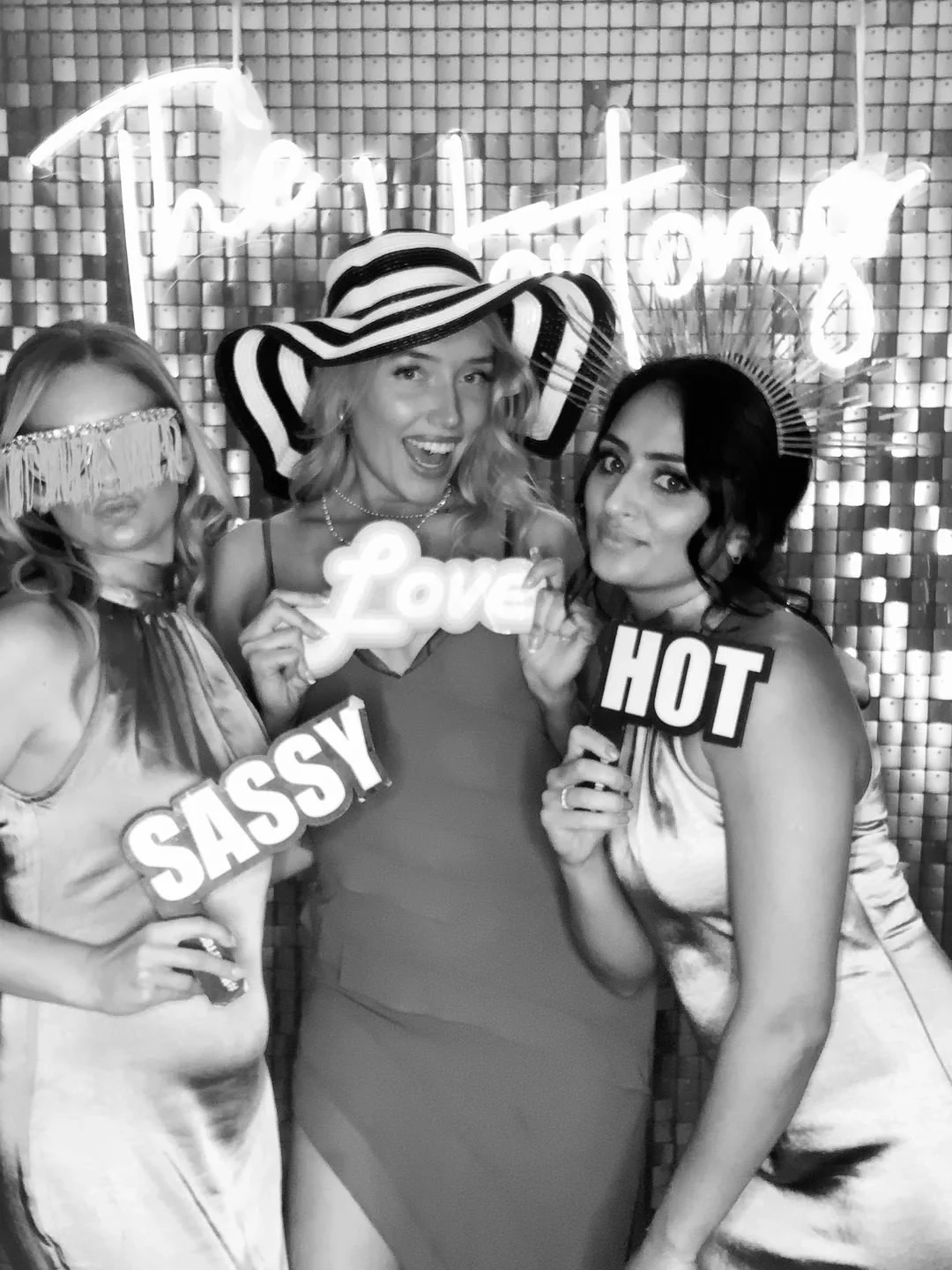Three women at a photo booth, holding signs that read 'Love', 'Sassy', and 'Hot', with a neon sign in the background, one woman wearing a large striped sunhat and a fringed eye mask, all dressed stylishly.