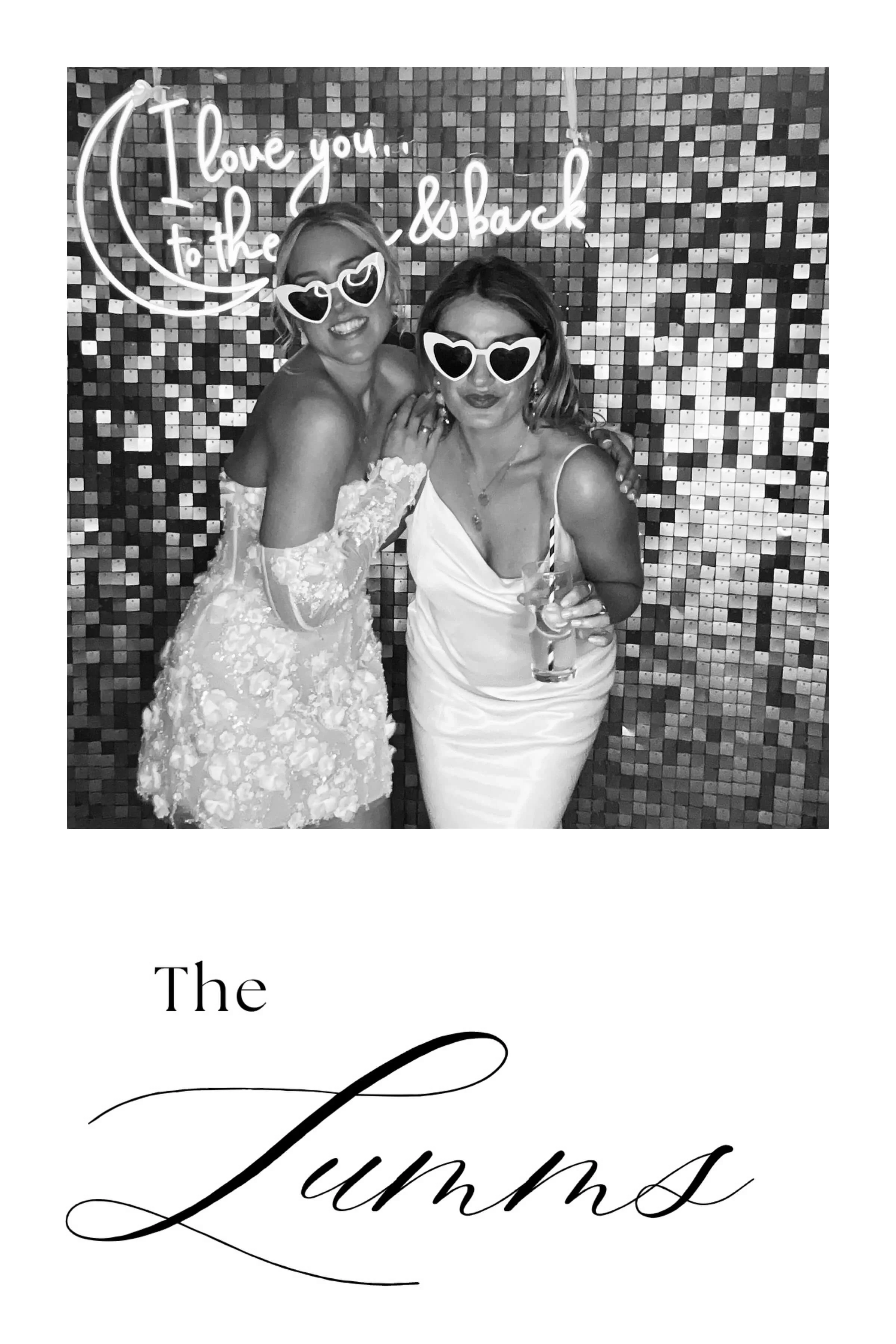 Black and white photo of two women wearing sunglasses, one in a lace dress and the other in a satin dress, standing in front of a mosaic wall with neon sign saying 'I love you to the moon and back.'