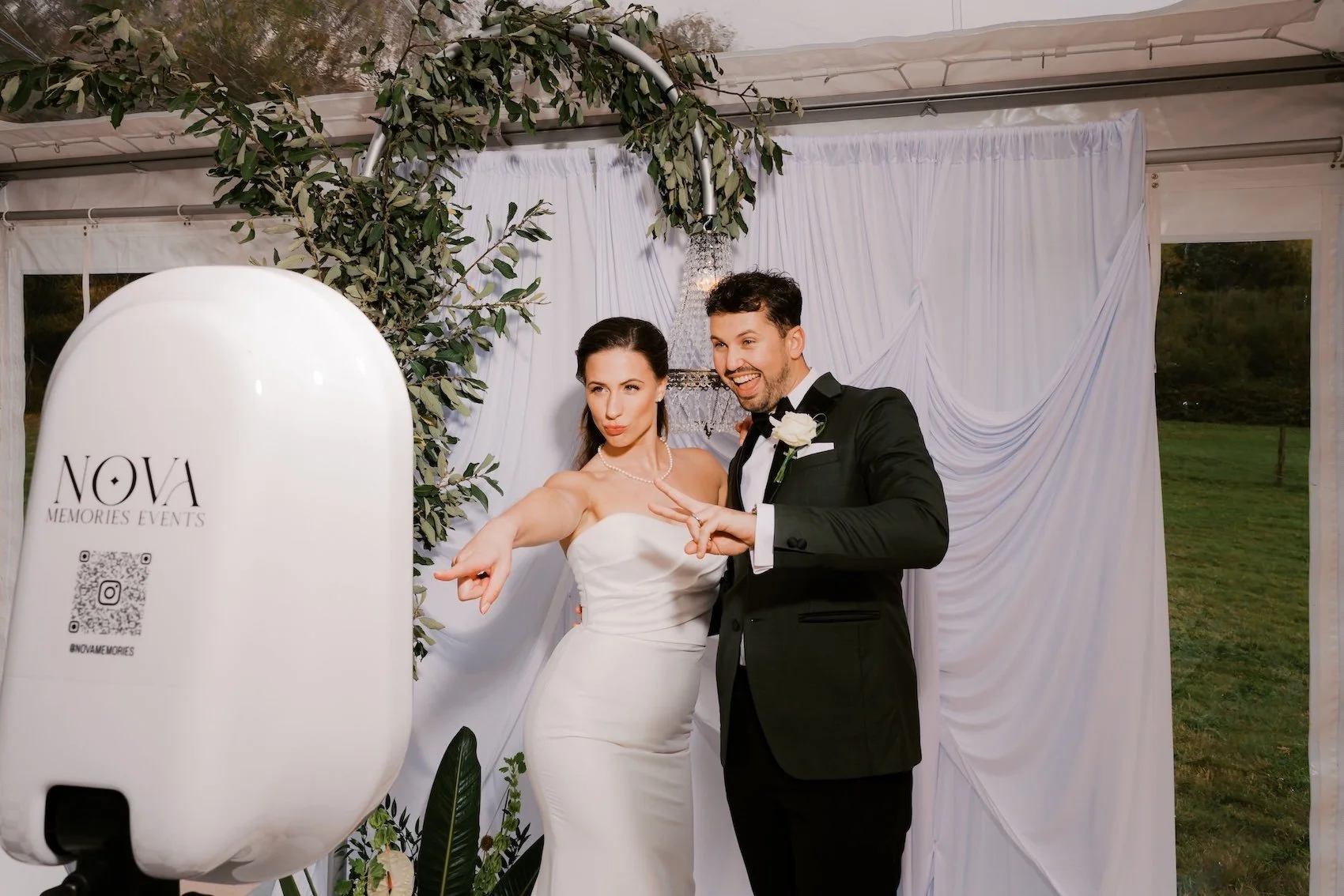 A bride and groom pose playfully during their wedding reception under a white draped backdrop with greenery and a chandelier, outside in a grassy area.