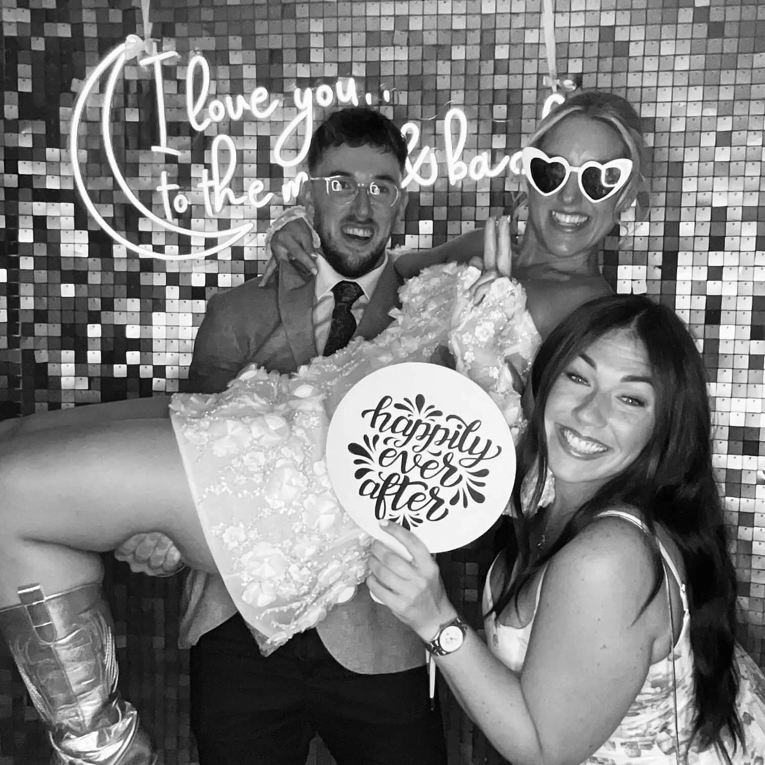 A group of four friends, including a bride and groom, celebrating at a wedding reception. The bride is holding a sign that reads 'happily ever after,' and the groom is holding a bouquet of flowers. They are posing in front of a decorative mirrored wa