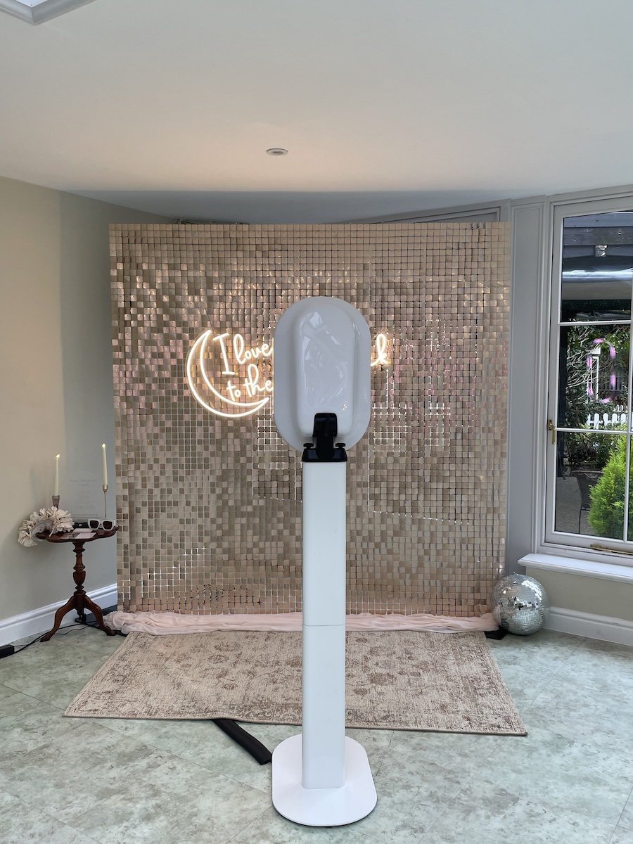 a decorative pink sequin backdrop with a neon sign that reads 'I love you to the moon and back'. There is a small wooden table with candles and flowers on the left and a disco ball on the right near a window on the right side of the room.