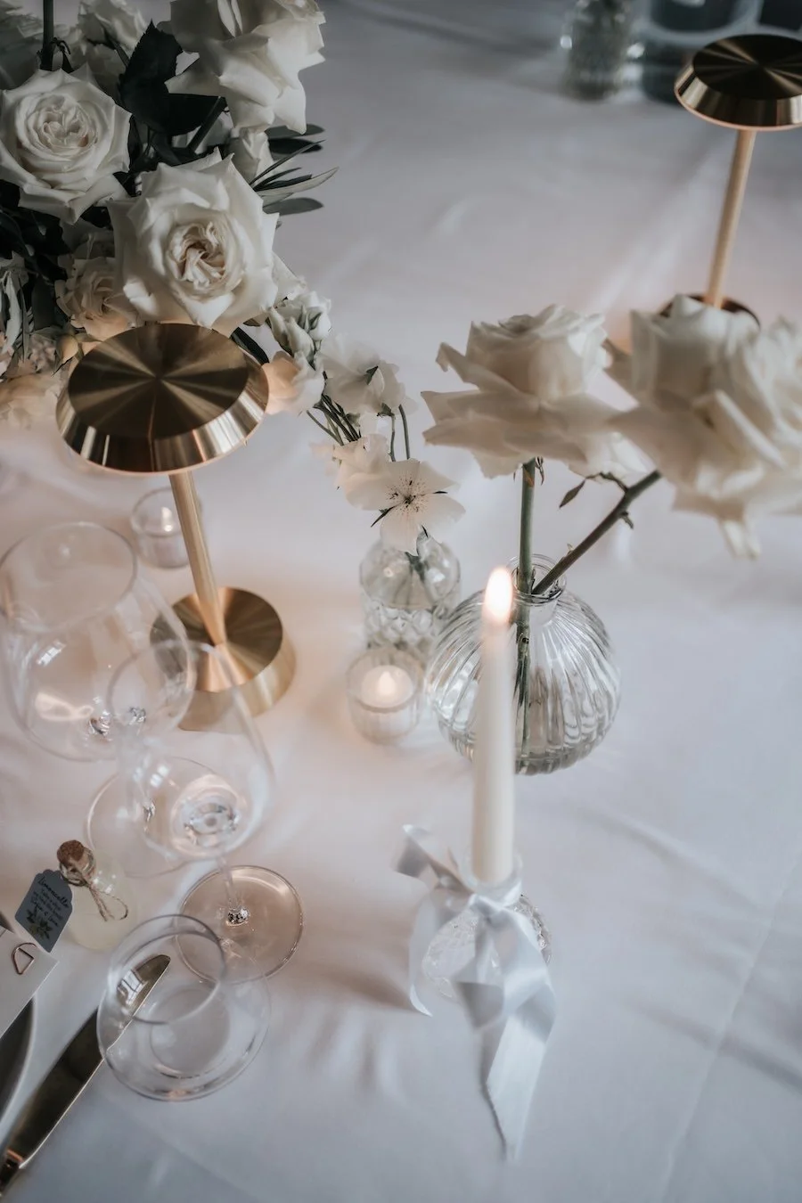 A table centerpiece with white roses, glass vases, a tall white candle with a white ribbon, and two modern gold lamps, set on a white tablecloth.