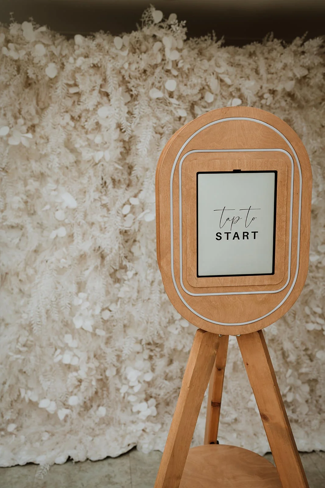A  vintage style photo booth a digital screen displaying 'tap to START' against a background of white, textured, floral wall.