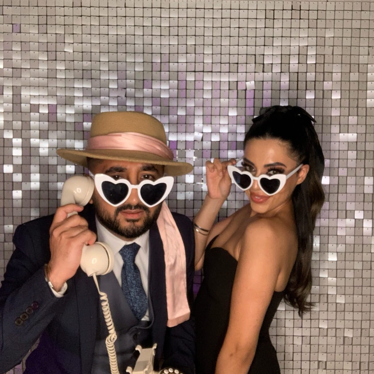 Two people at a party wearing heart-shaped sunglasses, with a woman in a black strapless dress and a man in a dark suit and tie, holding a vintage rotary phone, standing in front of a reflective, mosaic wall.