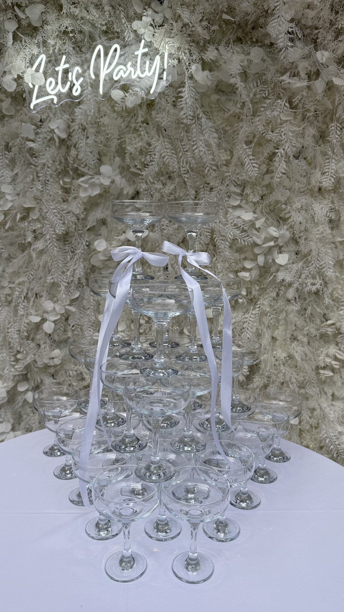 A pyramid of champagne glasses decorated with white ribbons, set against a backdrop of white floral and foliage decorations with a neon sign that reads 'Let's Party!'.