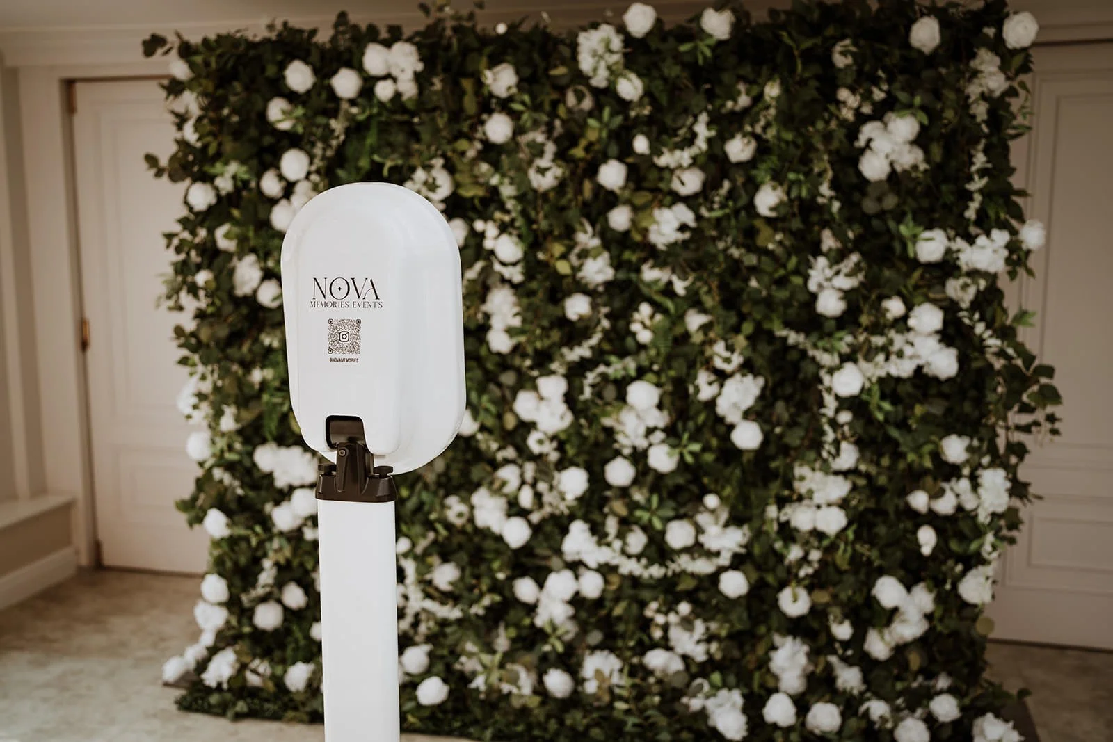 Photo booth in front of a wall decorated with white flowers. The dispenser has the words Nano Memories Events written on it.