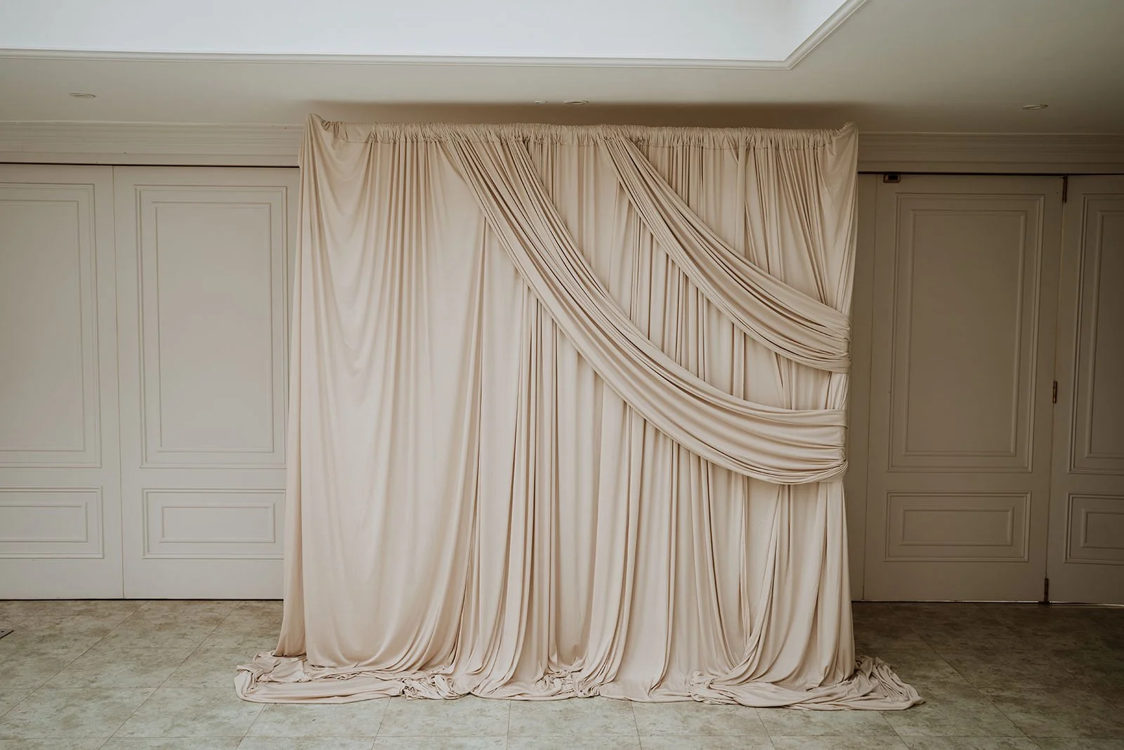 A large cream-colored draped backdrop  draped folds hangs against a wall, possibly for a wedding or event backdrop, in a room with white paneled walls and tiled flooring.