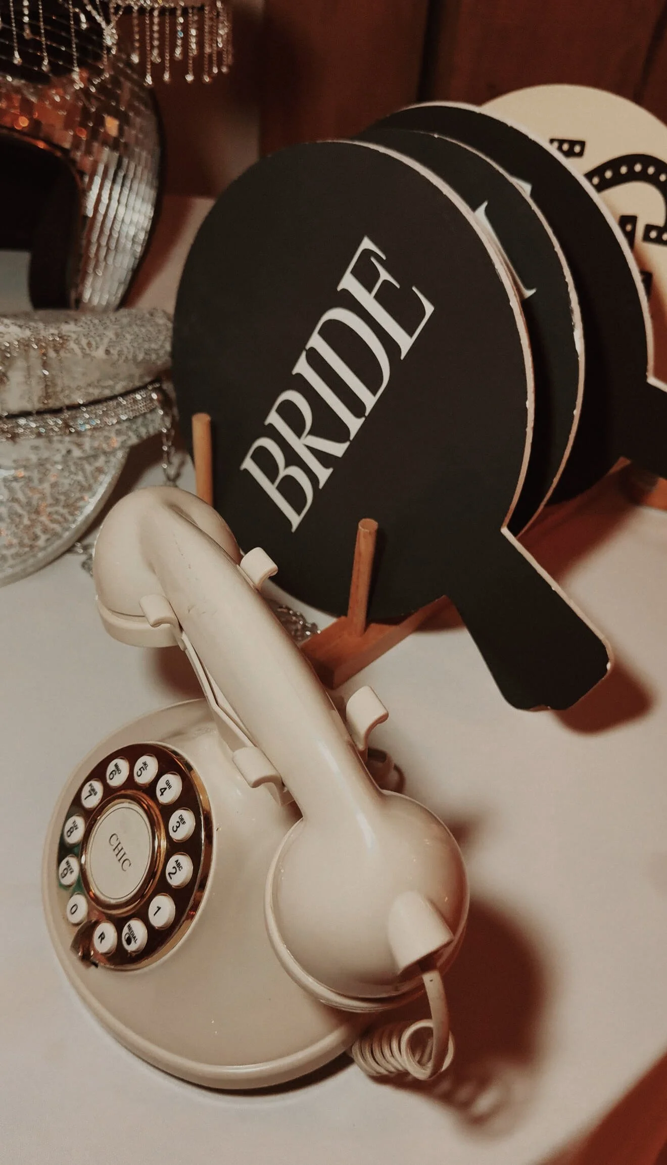 Vintage cream-colored rotary telephone with a cord, placed on a white surface. Behind it, there is a black circular sign with the word 'BRADE' written in white, held by small wooden stands. The background includes a sparkly silver decorative item and