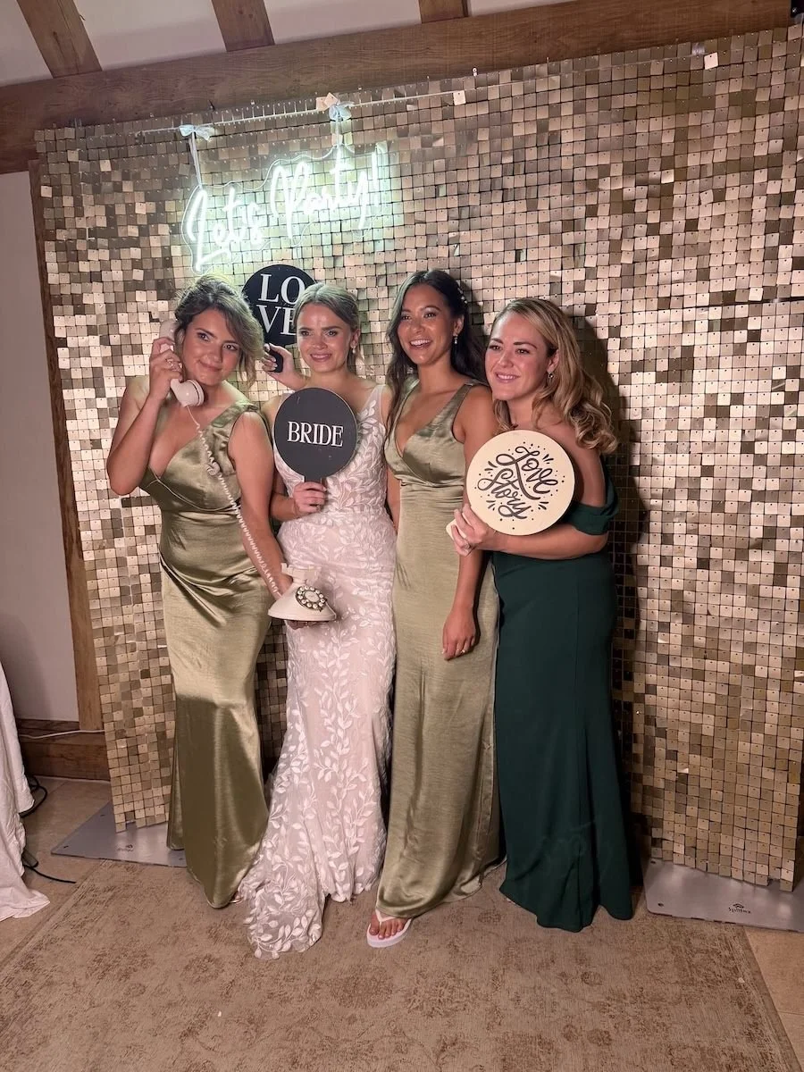 Four women dressed in formal gowns at a wedding, holding signs that say 'LOVE', 'BRIDE', and 'Love & Happy'. They are standing in front of a gold mosaic backdrop with a neon sign that says 'Let’s Party'.