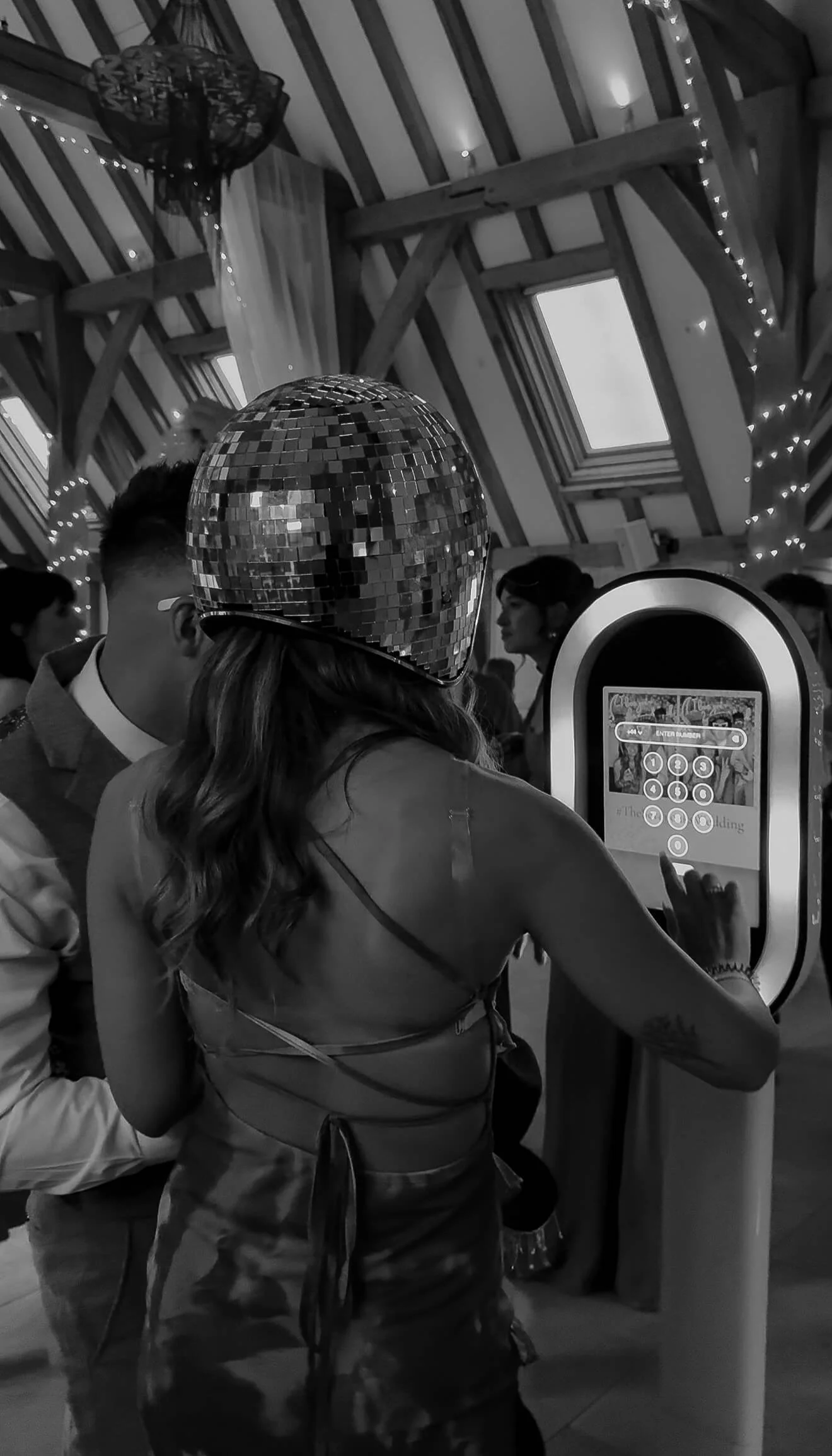 A woman wearing a shiny disco ball hat and a strappy top is using a touchscreen jukebox in a party setting. Other guests are visible in the background under a wooden ceiling with string lights.