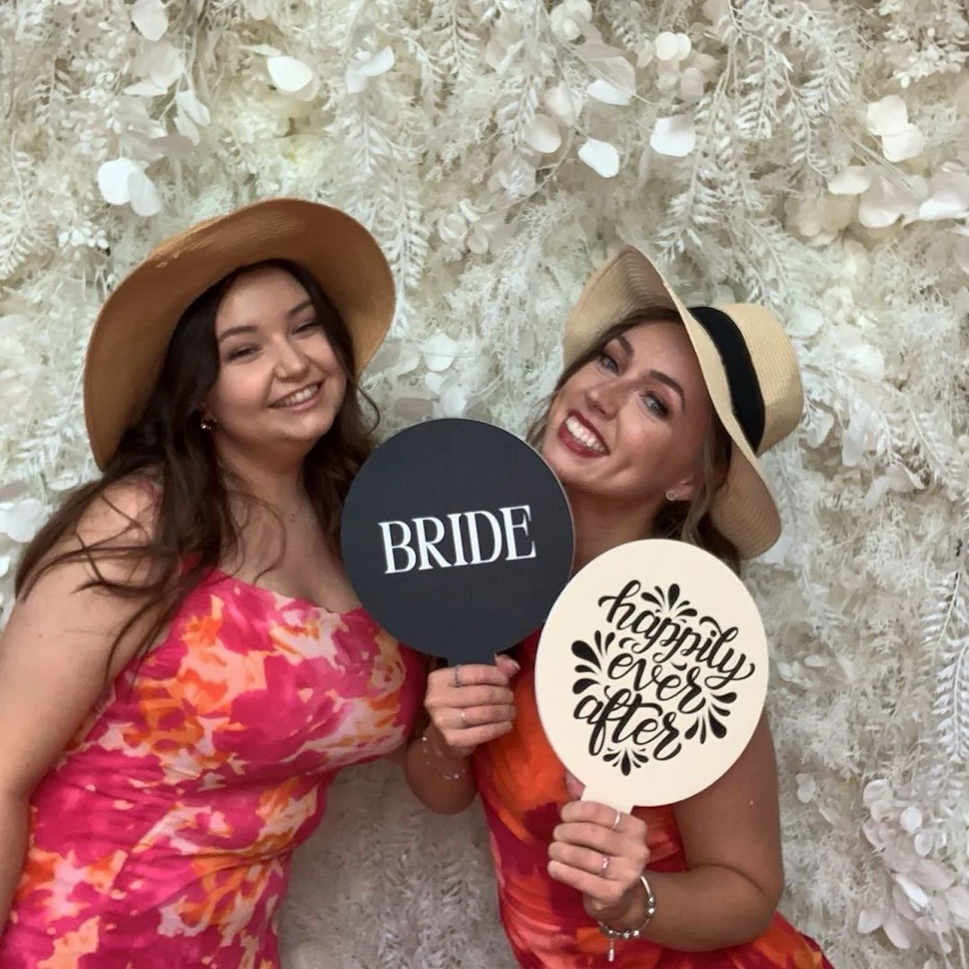 Two women at a wedding reception, one wearing a pink floral dress and a wide-brimmed hat, the other in a red dress with a black hat. They are holding signs that say 'BRIDE' and 'happily ever after' respectively, standing against a white floral backdr