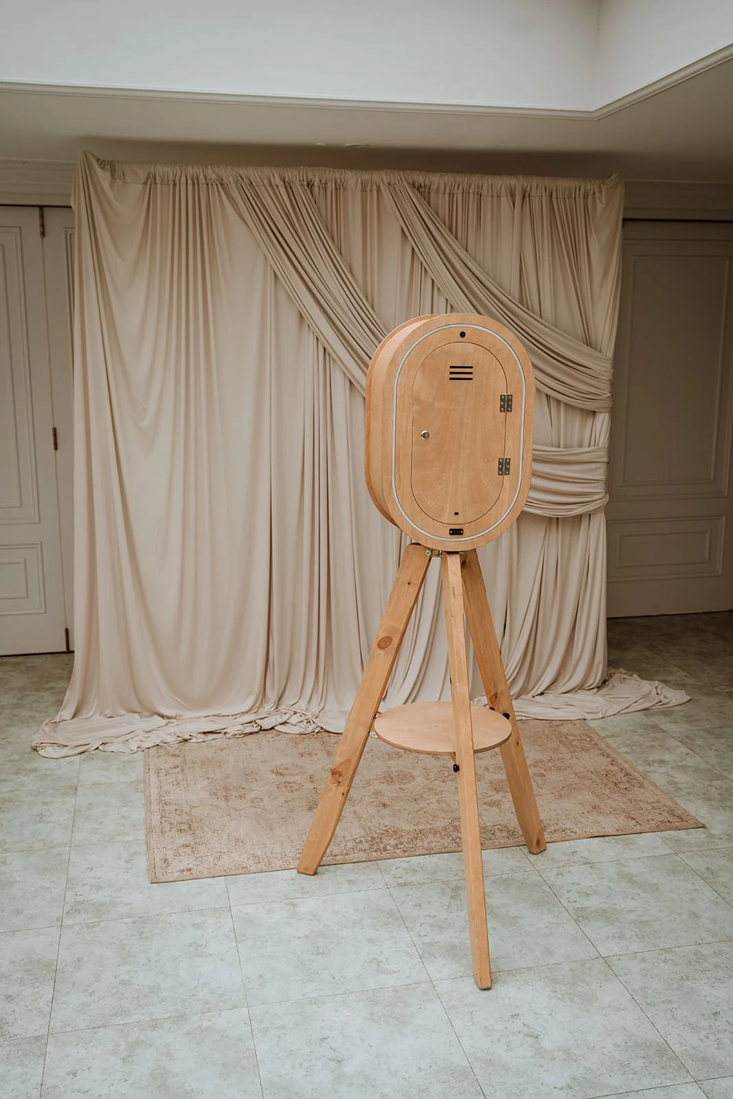 Uncovered wooden photo booth on tripod stand in front of beige draped curtain backdrop in an indoor setting.