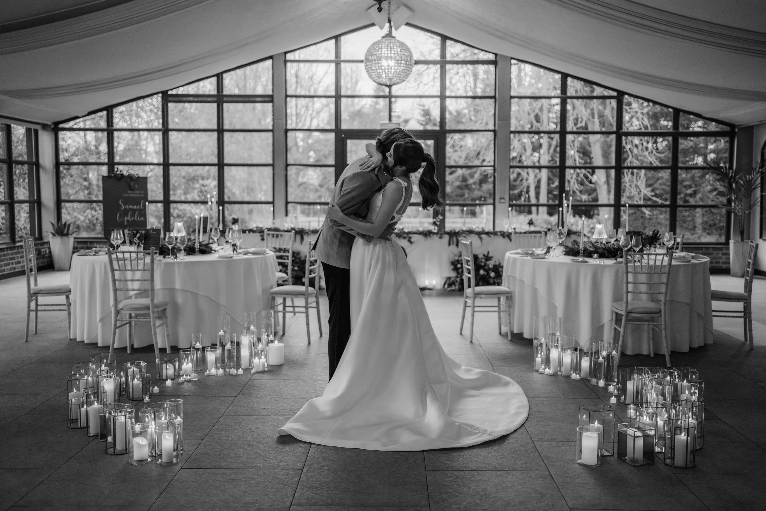 A couple sharing a kiss at their wedding reception in a decorated indoor venue with large windows, hanging light fixture, round tables with tableware, and candles on the floor.