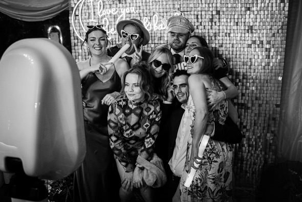 Group of women and one man taking a fun selfie at a party, with some wearing sunglasses and holding photo booth props, and a glittery backdrop with a neon sign.