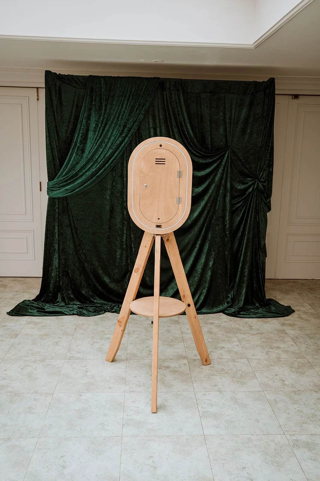 A wooden photo booth with a black velvet curtain backdrop on a tiled floor.