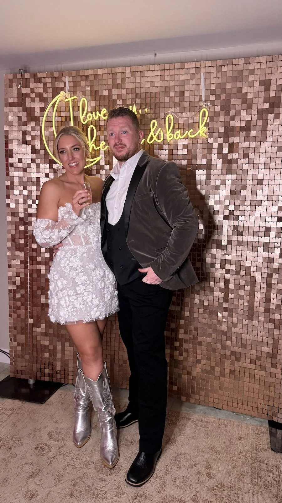 A couple dressed in formal attire standing in front of a sparkly backdrop with a neon sign that reads 'love it when you come back.' The woman is holding a drink, wearing a white, floral, off-the-shoulder dress with silver cowboy boots. The man is in 