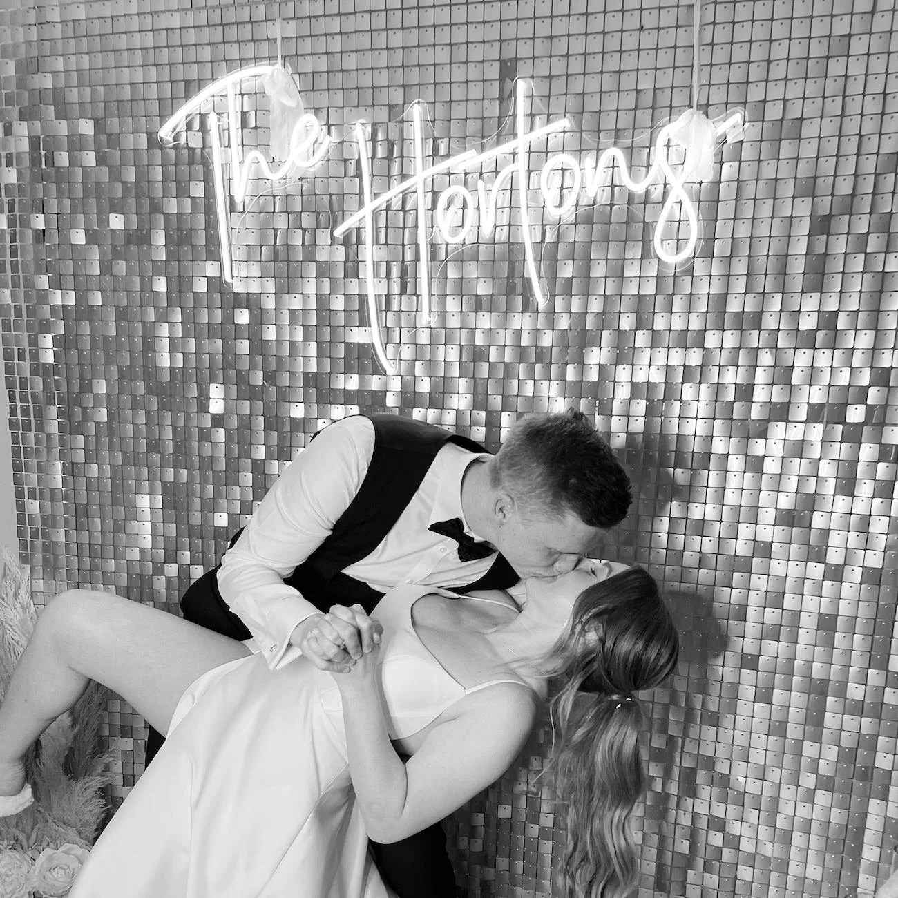 A couple sharing a kiss at their wedding, with a man in a tuxedo and a woman in a wedding dress, in front of a tiled wall with a neon sign reading 'The Knot'.