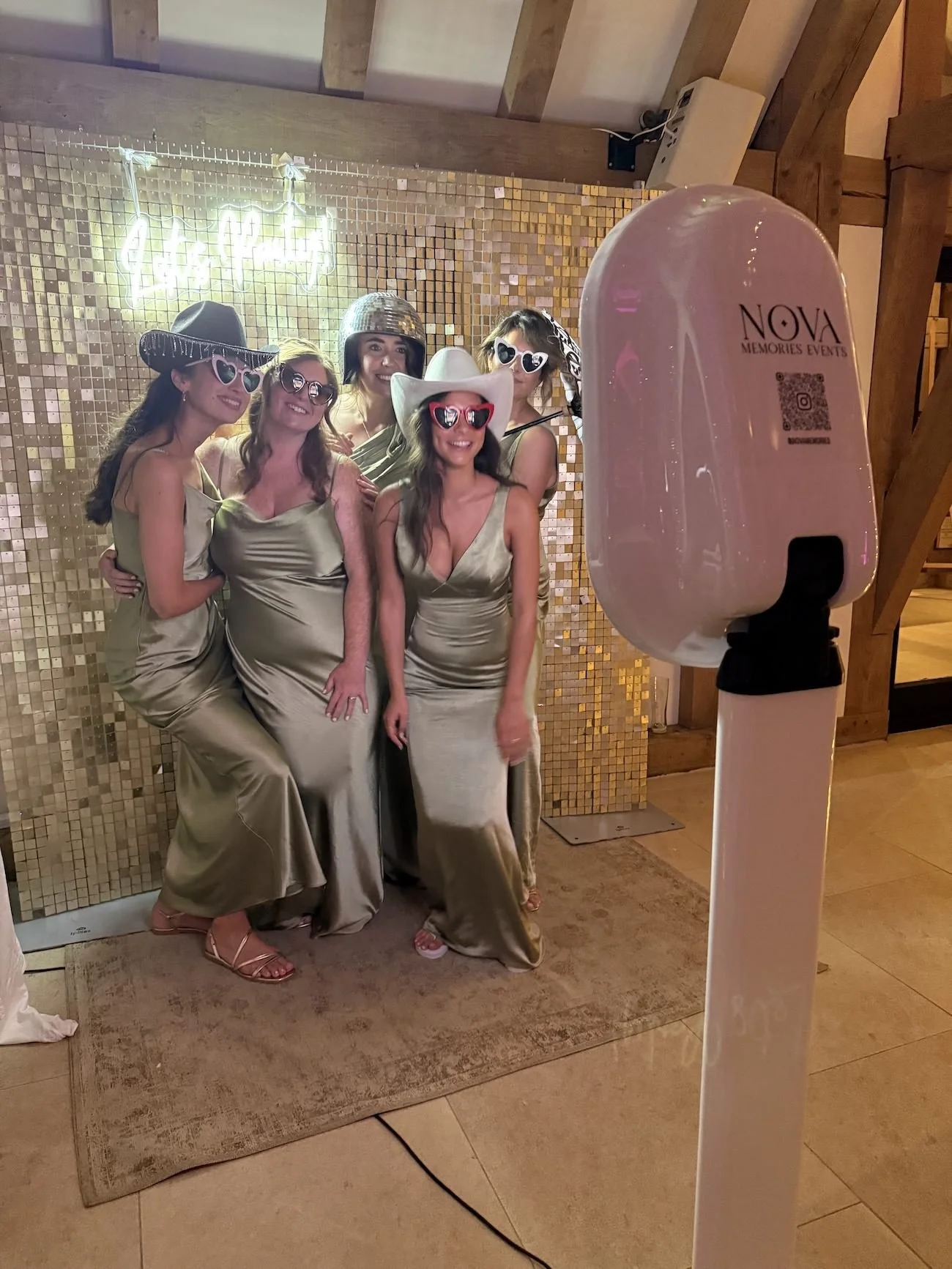 Group of five women dressed in gold satin gowns posing for a photo at a celebration, wearing various fun hats and oversized heart-shaped sunglasses, with a gold mosaic backdrop and a neon sign that says 'Happy Birthday'.