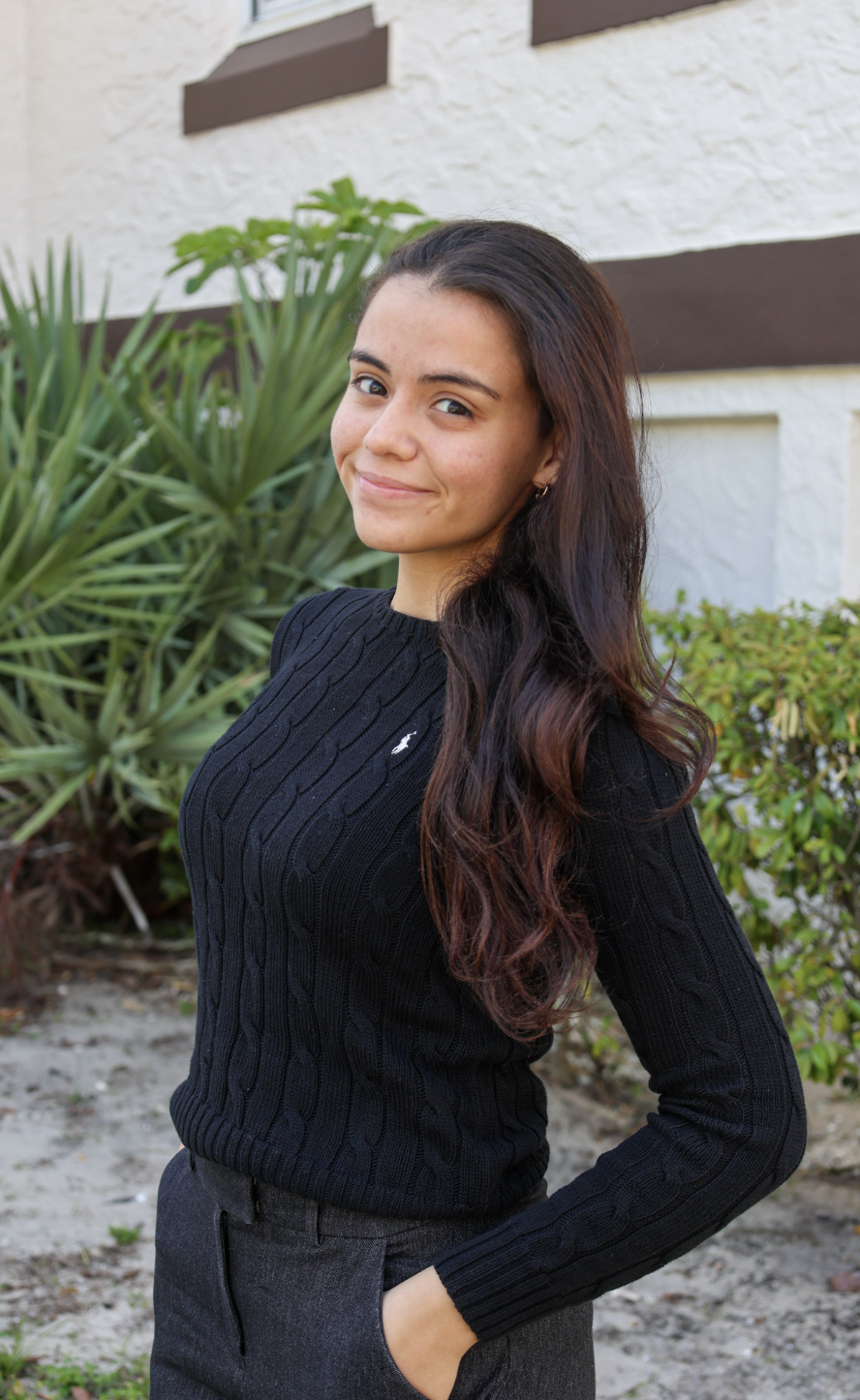 Young woman with long dark hair wearing a black cable-knit sweater and dark pants, smiling outdoors with greenery and a white building in the background.