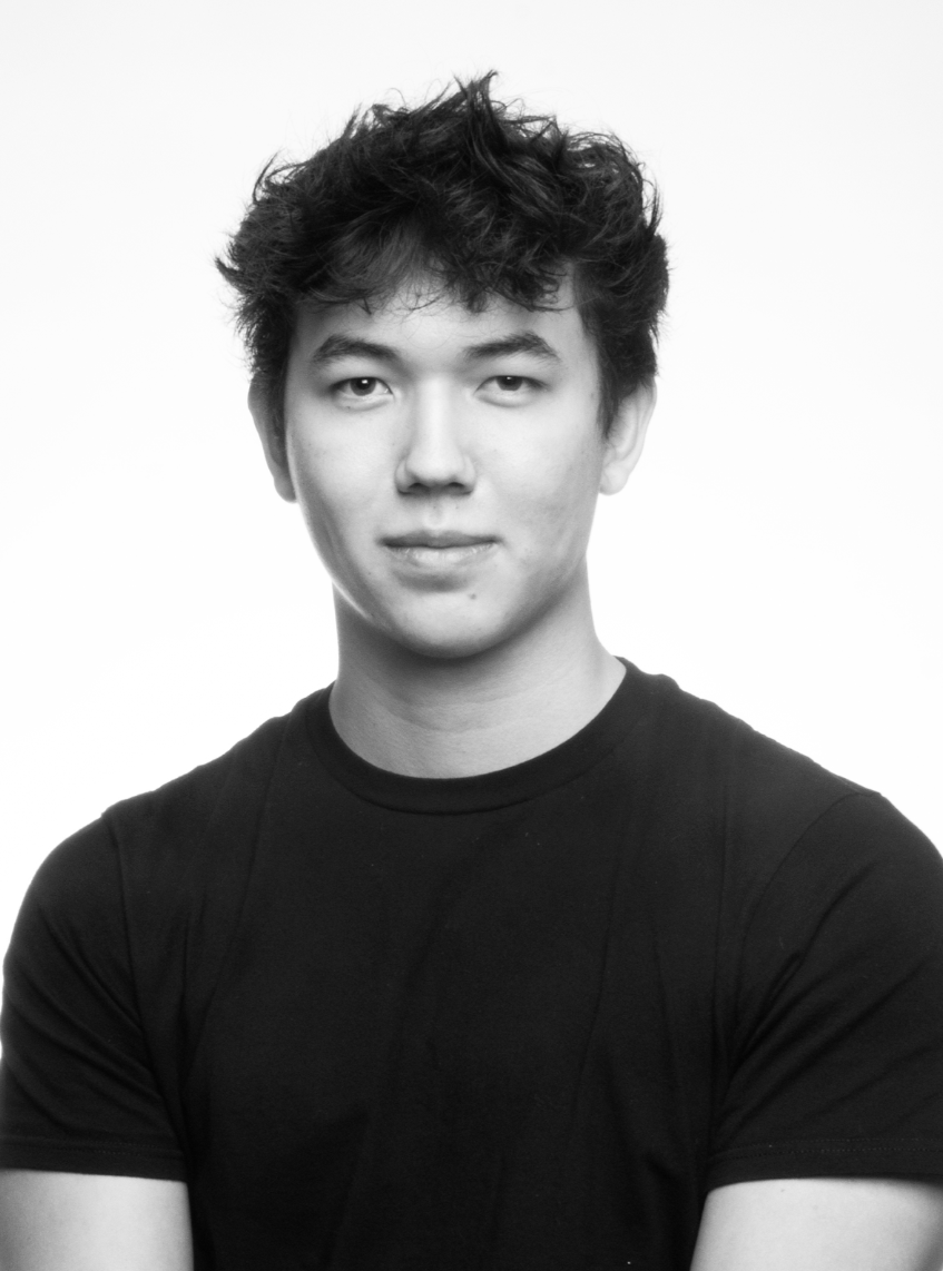 Black and white portrait of a young man with curly, tousled hair, wearing a plain black t-shirt, looking at the camera with a slight smile.