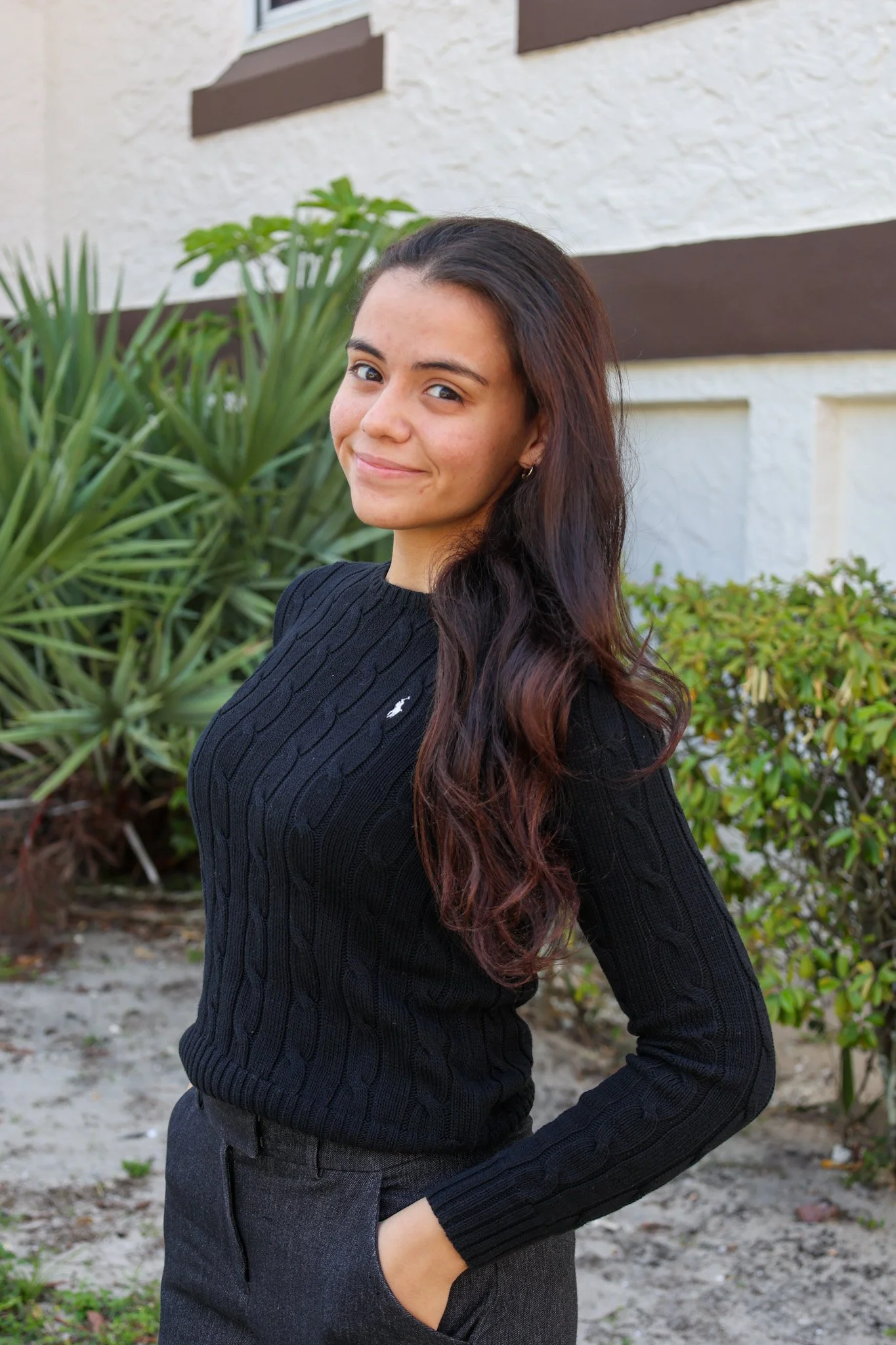 A woman with long, wavy dark hair and a slight smile, standing outdoors in front of green plants and a building with a white and brown exterior. She is wearing a black, ribbed sweater and dark pants.