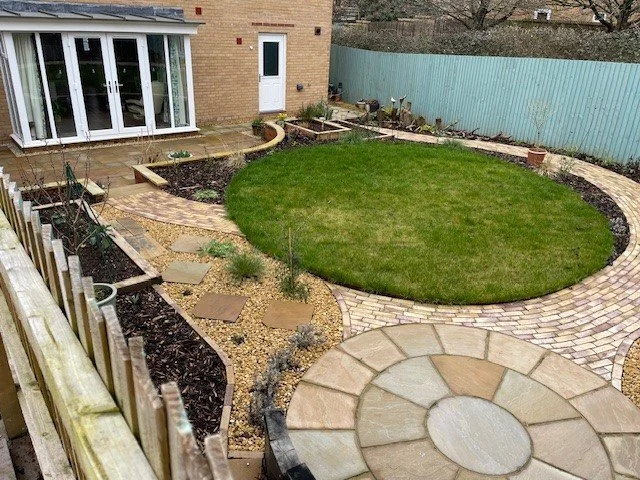Backyard garden with a circular lawn, paved pathways, raised garden beds, and a brick house with glass doors.