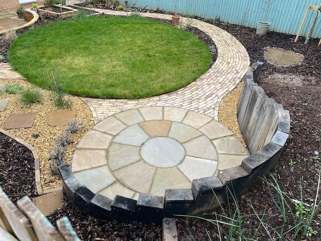 A circular garden with a green lawn, bordered by brick and stone pathways, and a curved retaining wall with a wooden fence in the background.