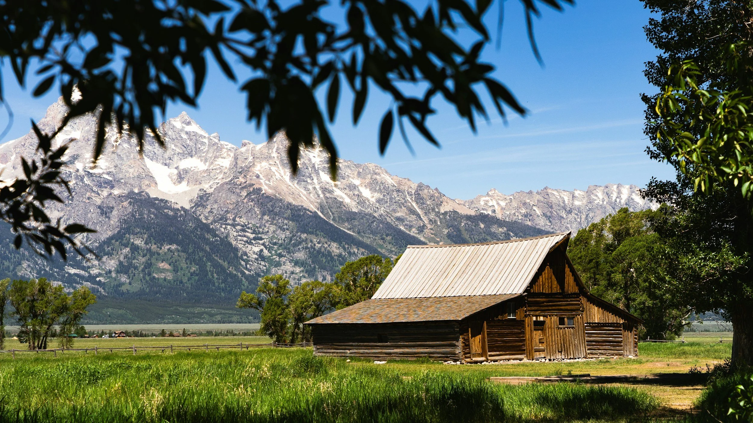 A rustic wooden cabin in a green meadow with mountains in the background and trees in the foreground.