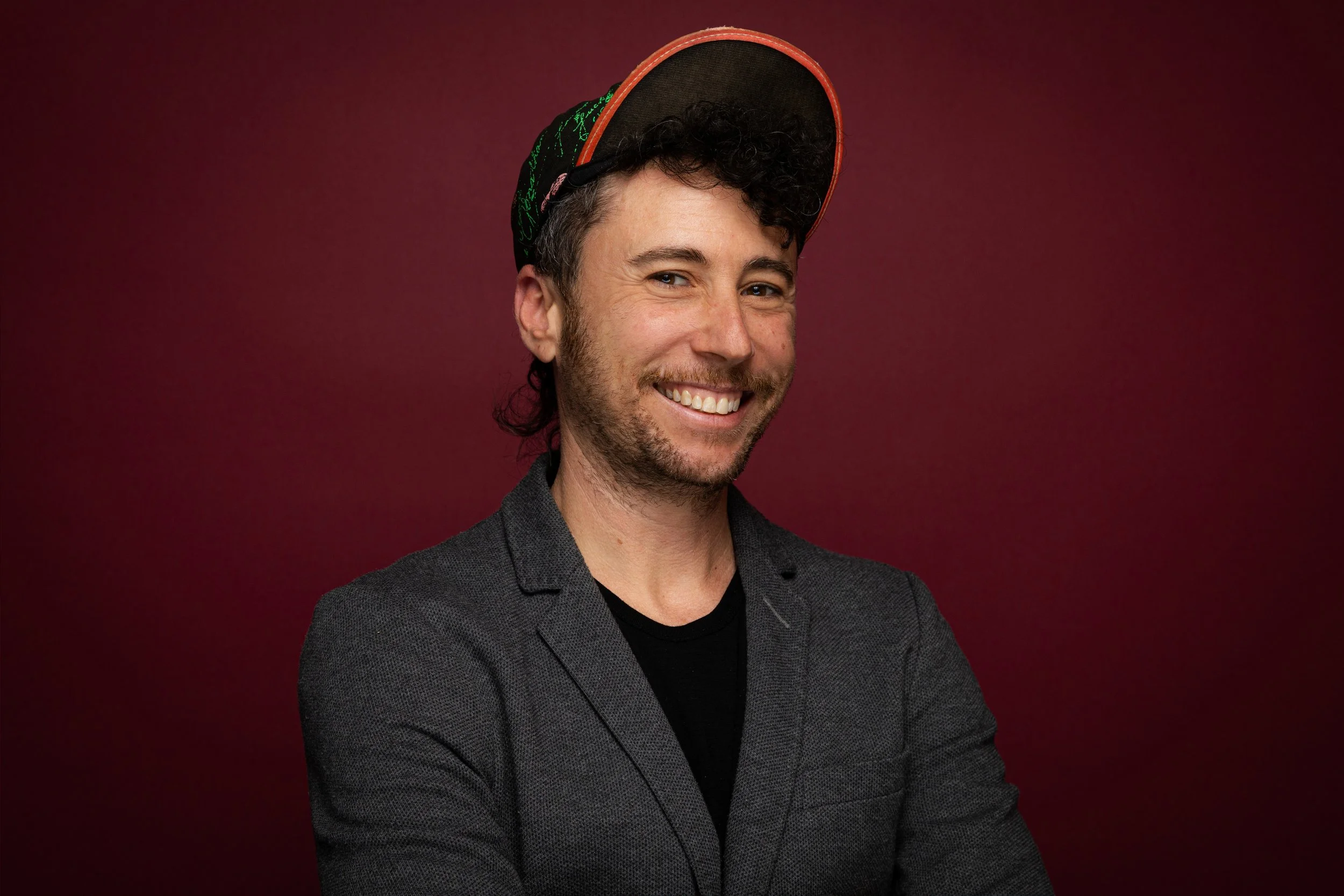 A smiling white transmasc person wearing a hat and a dark gray blazer poses in front of a maroon backdrop.