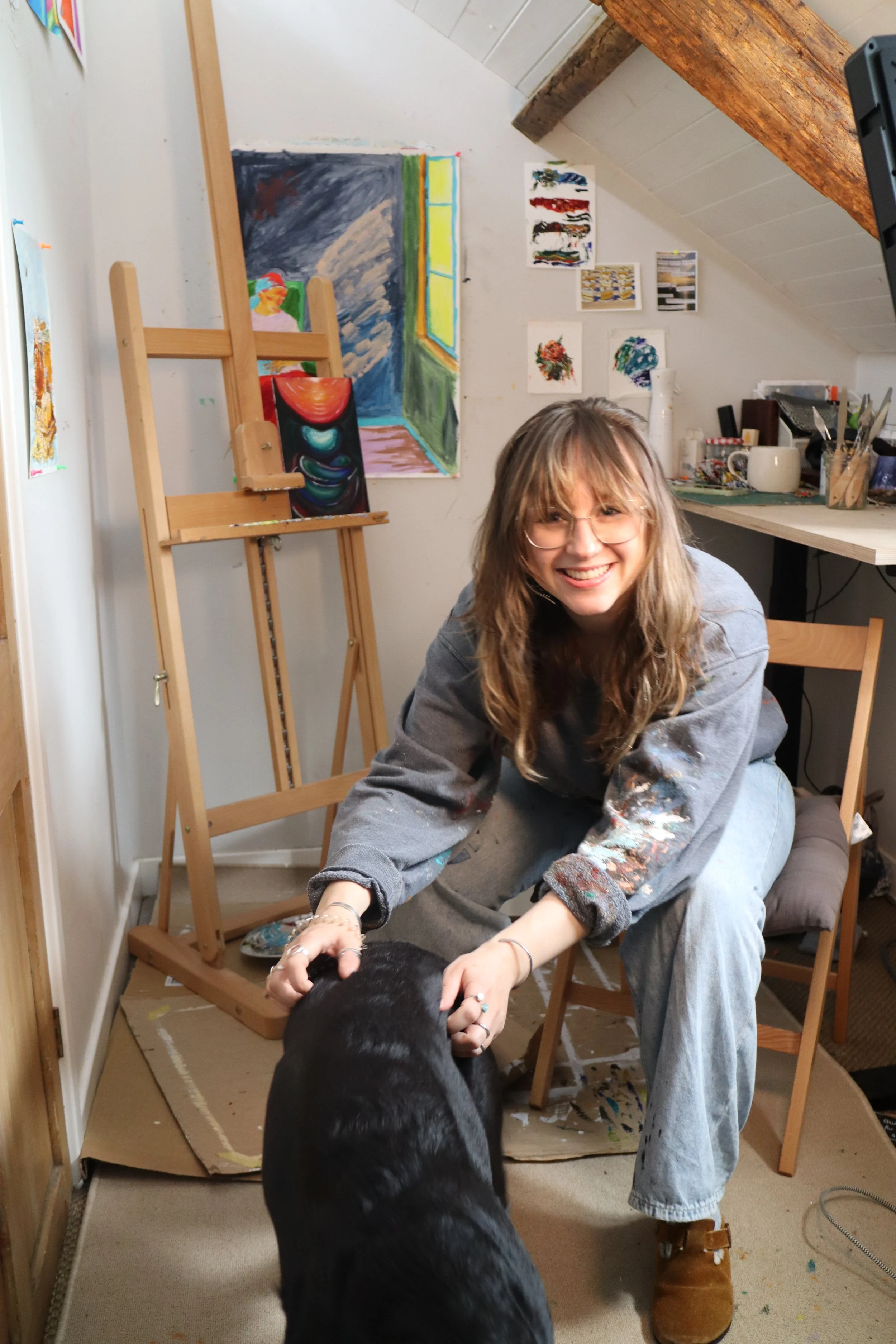 A woman smiling and petting a black dog in an art studio, with paintings on the wall and art supplies on a shelf in the background.