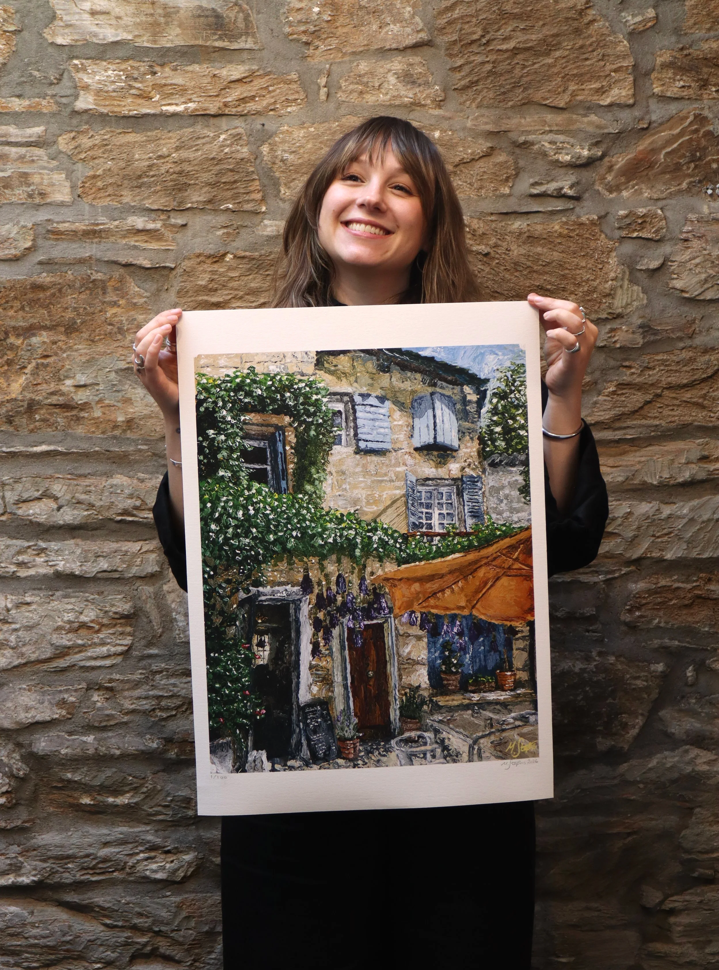 A woman smiling and holding a fine art print of a rustic house with a stone wall, window shutters, greenery, and a yellow umbrella, standing against a brick wall background.