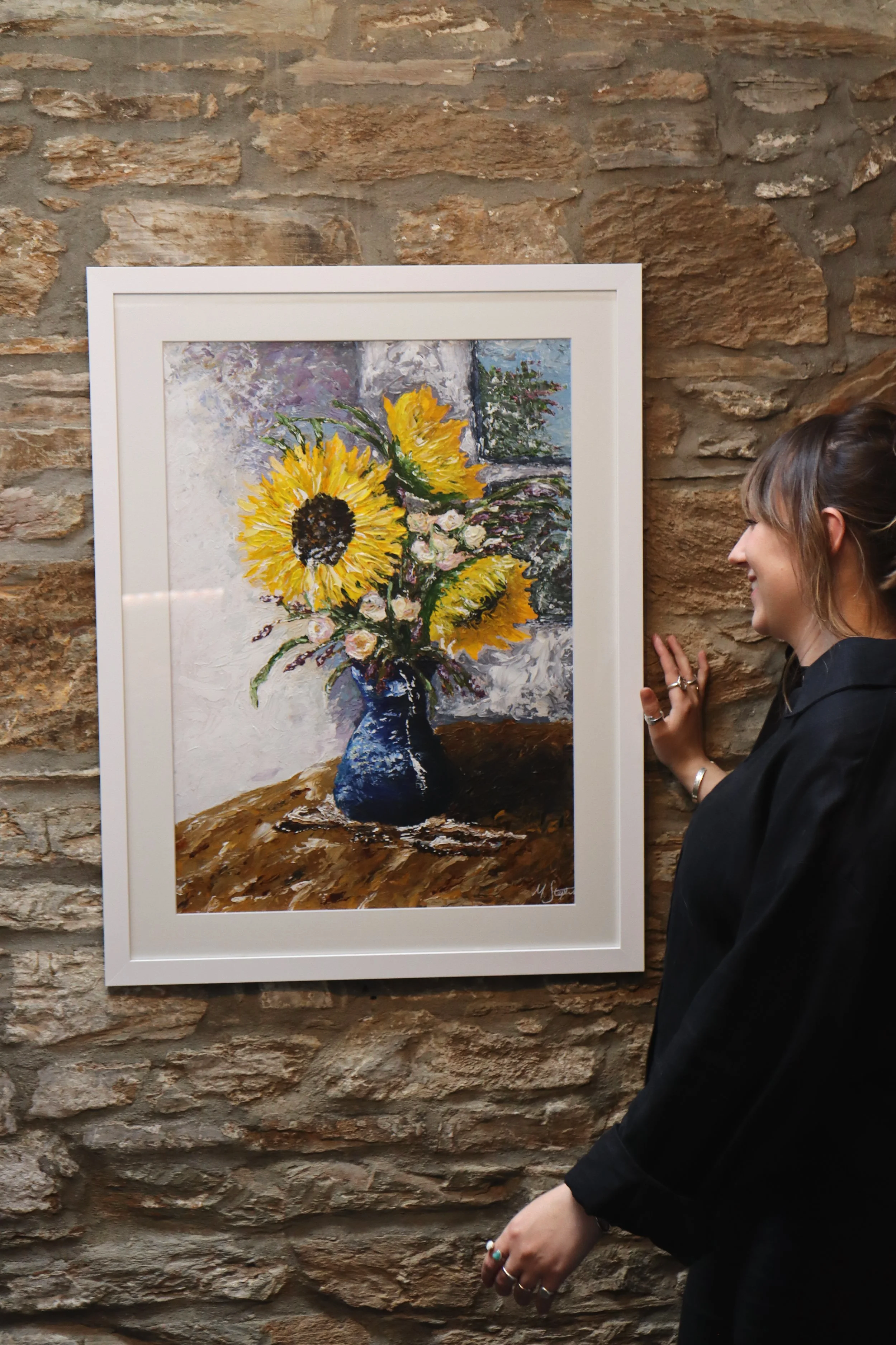 A woman in a black shirt smiling as she looks at a framed impressionist painting of sunflowers on a stone wall. The woman has her hair up and is wearing rings on her fingers.