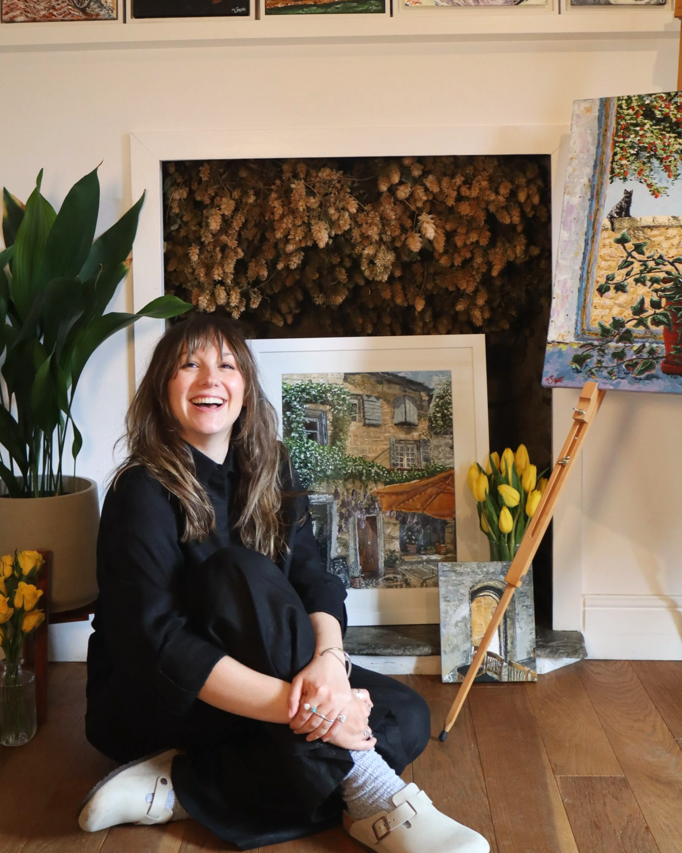 A young woman with long brown hair sitting on the floor, smiling and laughing, in an art studio, with paintings and artwork behind her, a large potted plant to her left, and a bouquet of yellow tulips on a table beside her.