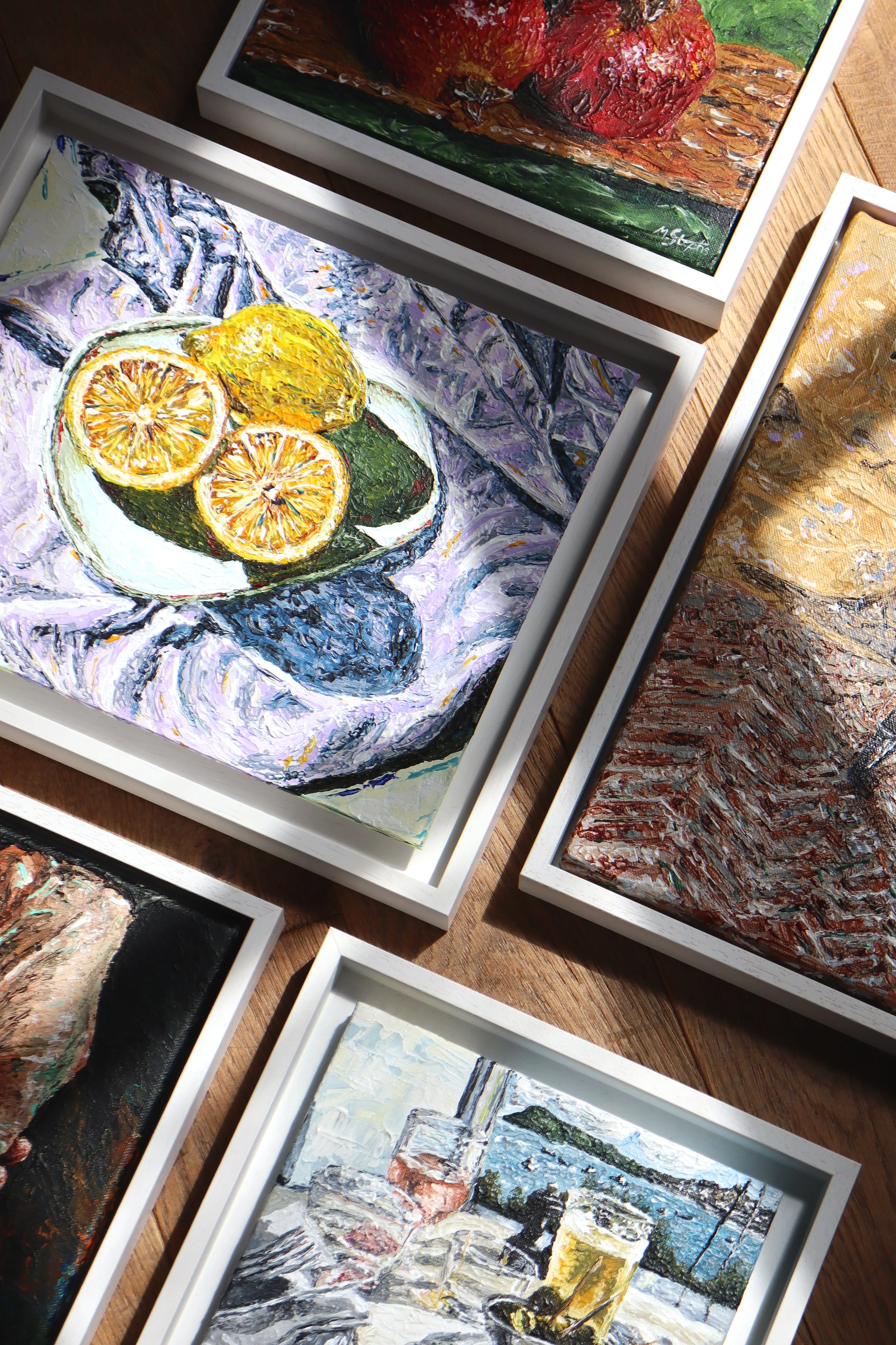 Paintings of various scenes, including still life with lemons, a table scape overlooking a harbour, and cityscape, framed in white and displayed on a wooden surface.