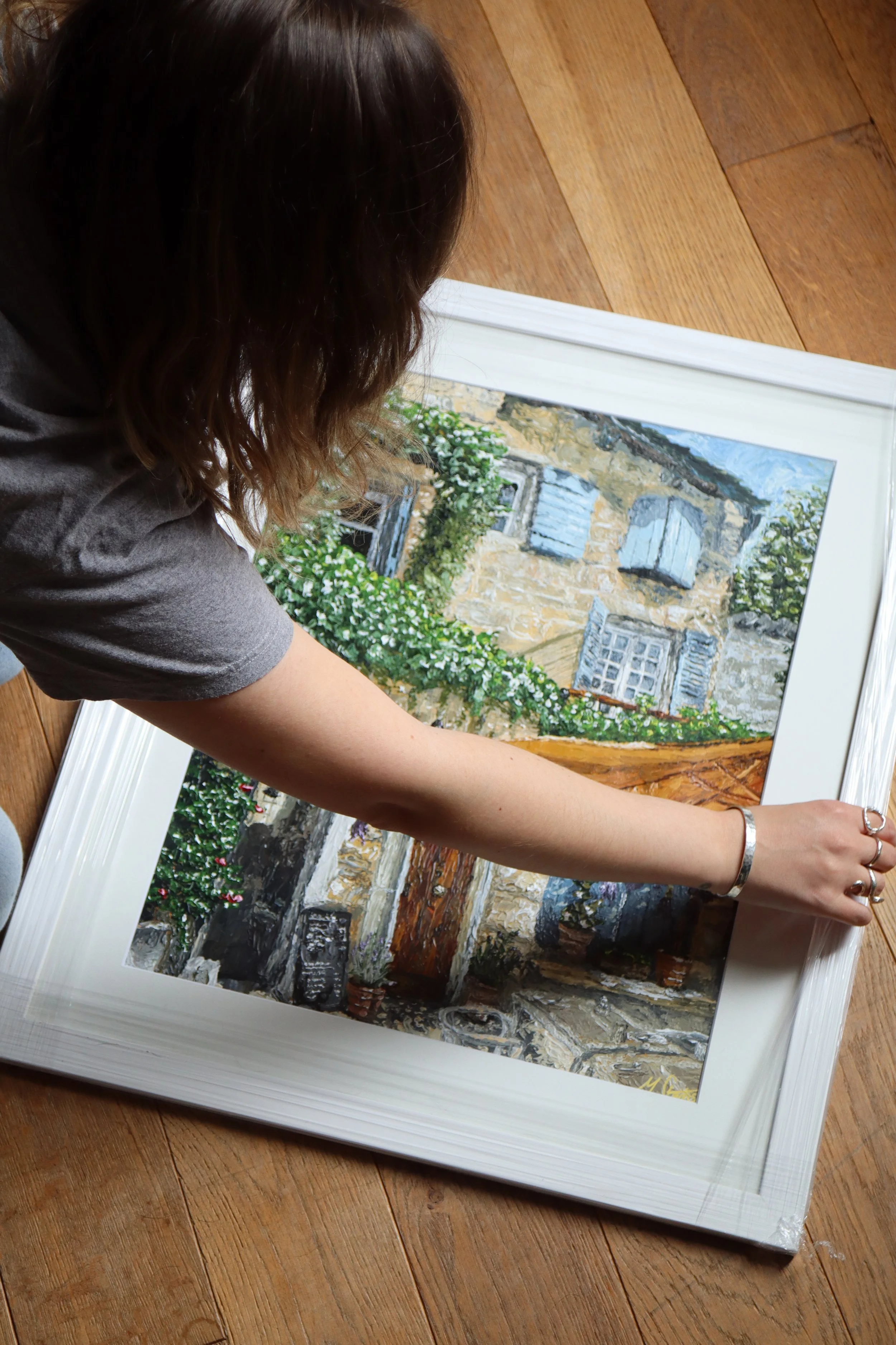 A person with brown hair leaning over a painting of a stone building with blue shutters and surrounding greenery on the floor.