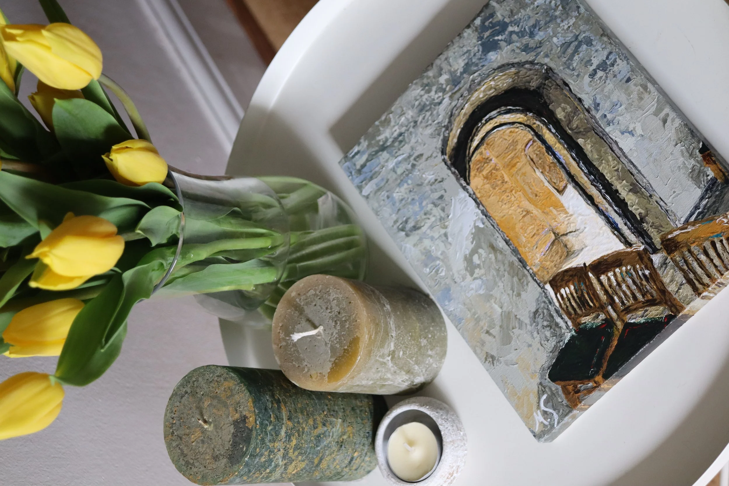 Yellow tulips in a glass vase with candles and impressionist painting of an old church archway on a white table.