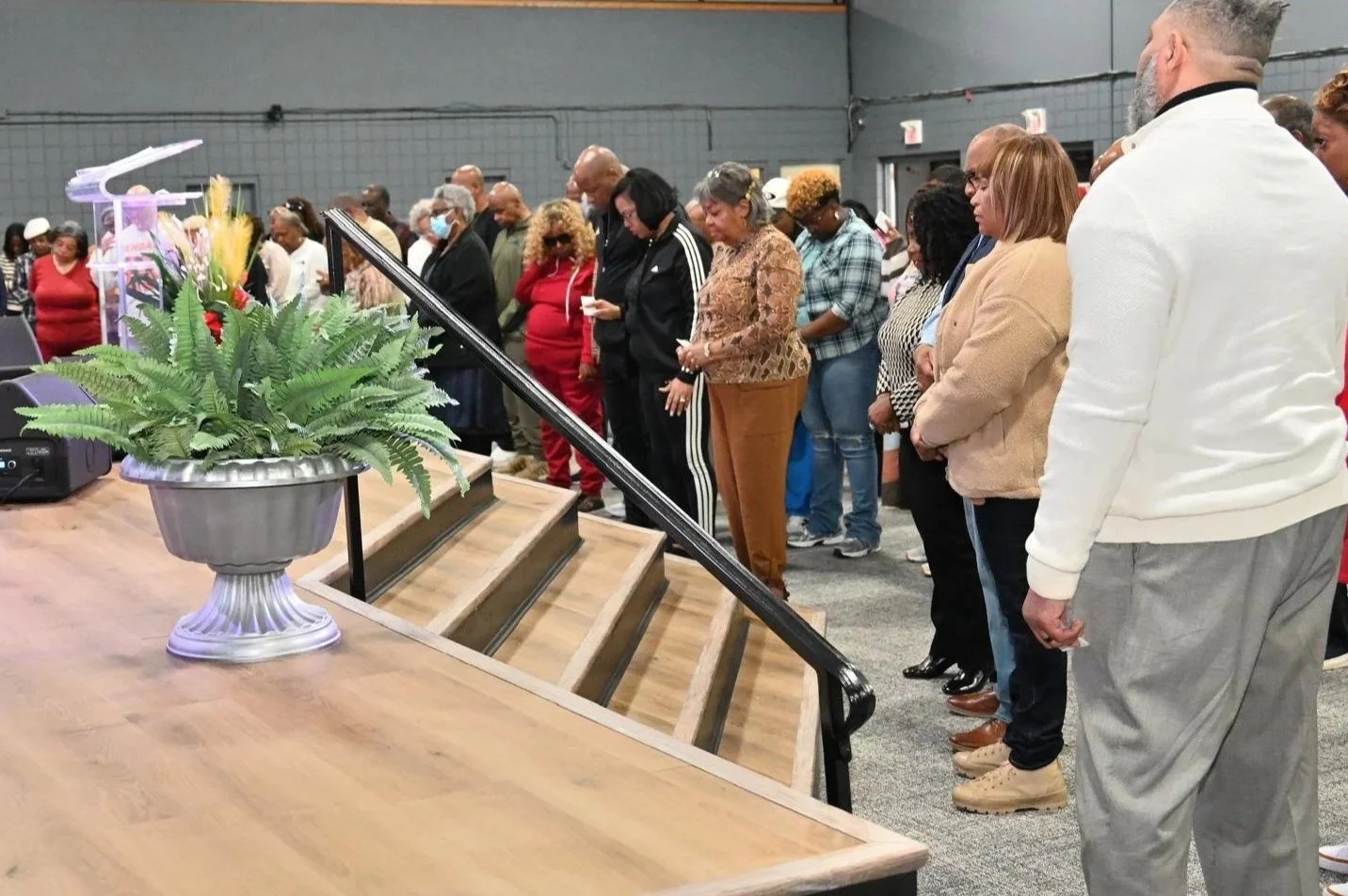 Group of people praying with their heads bowed, standing in a line inside a dark indoor space, near a wooden table with a large green fern plant in a silver pot.