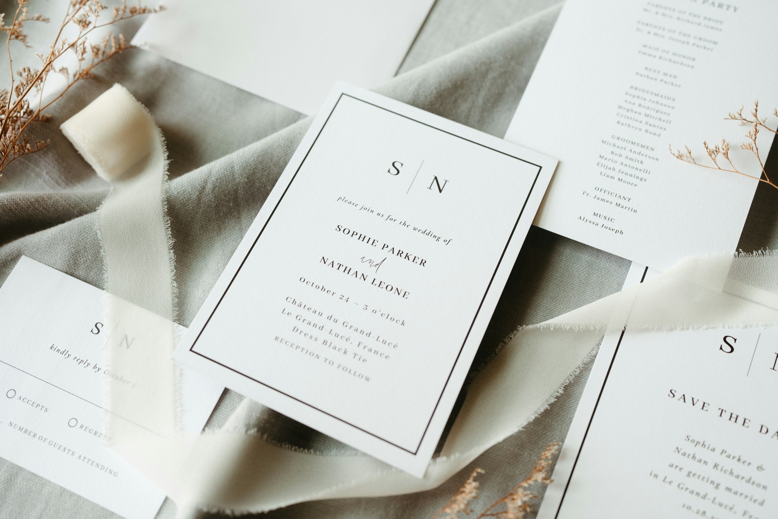 Close-up of wedding invitations and save-the-date cards with a neutral color palette, some cards on beige fabric, along with dried flowers and a ribbon.